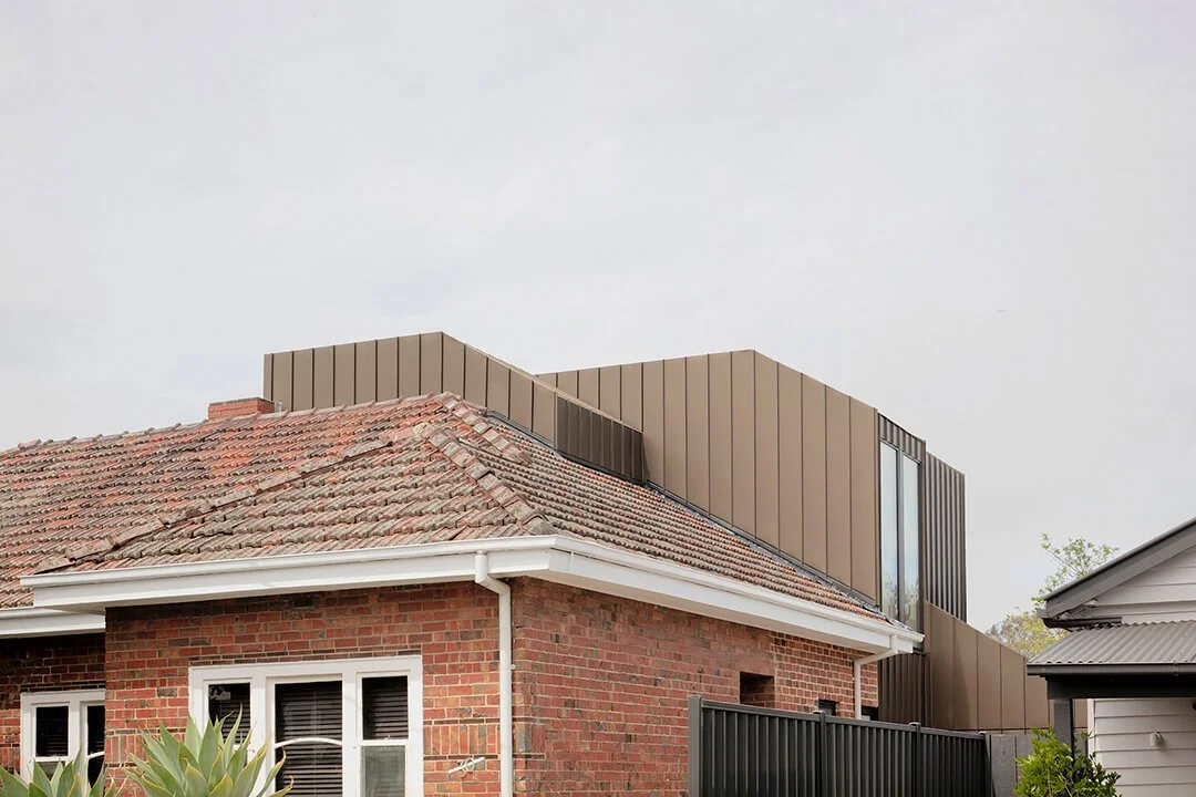 Contemporary metal-clad upper-level addition rising behind a red brick heritage home, contrasting old tiled roof with modern vertical cladding