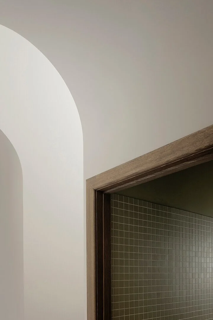 Minimalist interior featuring a white arched wall next to a wooden door frame. Behind the door, green mosaic tiles add subtle contrast. Calm ambiance.