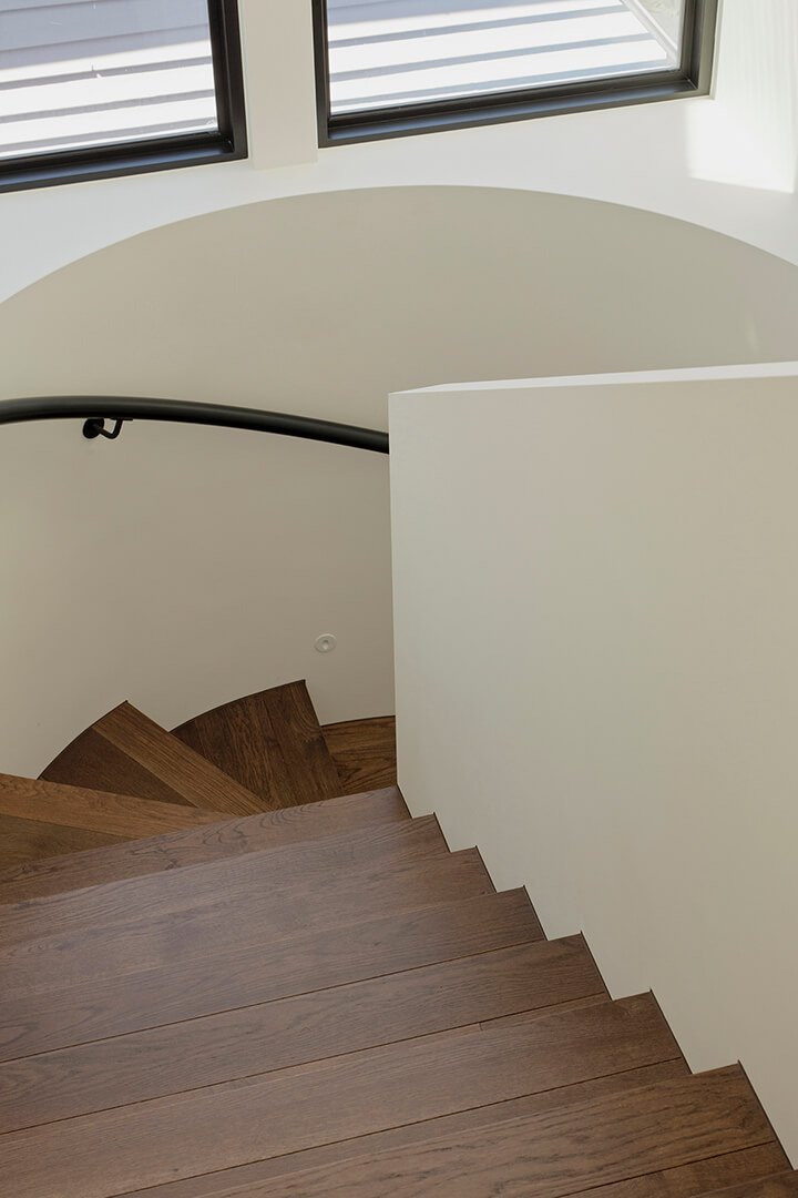 Spiral wooden staircase with sleek metal handrail, descending under a large, bright window. The setting feels modern and airy, with soft natural light.