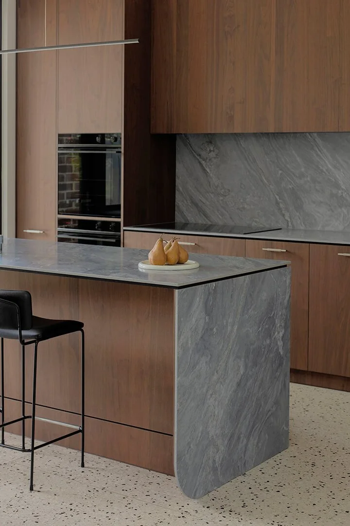 Modern kitchen with dark wood timber veneer cabinets, a grey marble backsplash, and a countertop. A minimalist decor add a sleek, calm vibe.