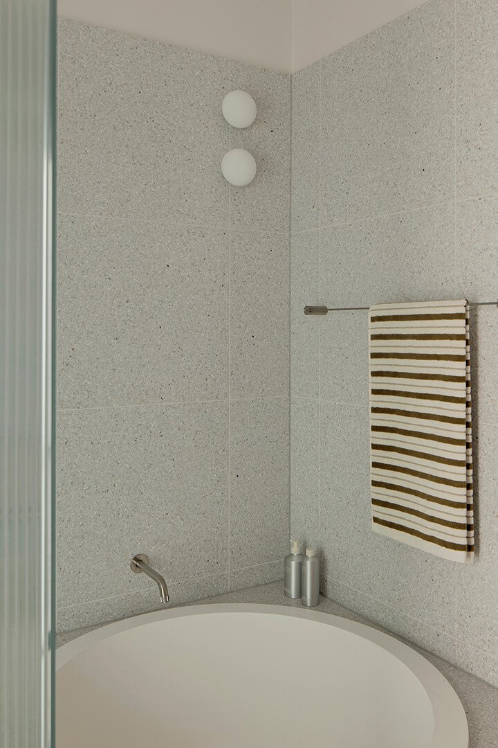 Minimalist bathroom with a round bathtub, grey tiled walls, and a feature towel rail. Two wall lights add a serene, modern ambience.