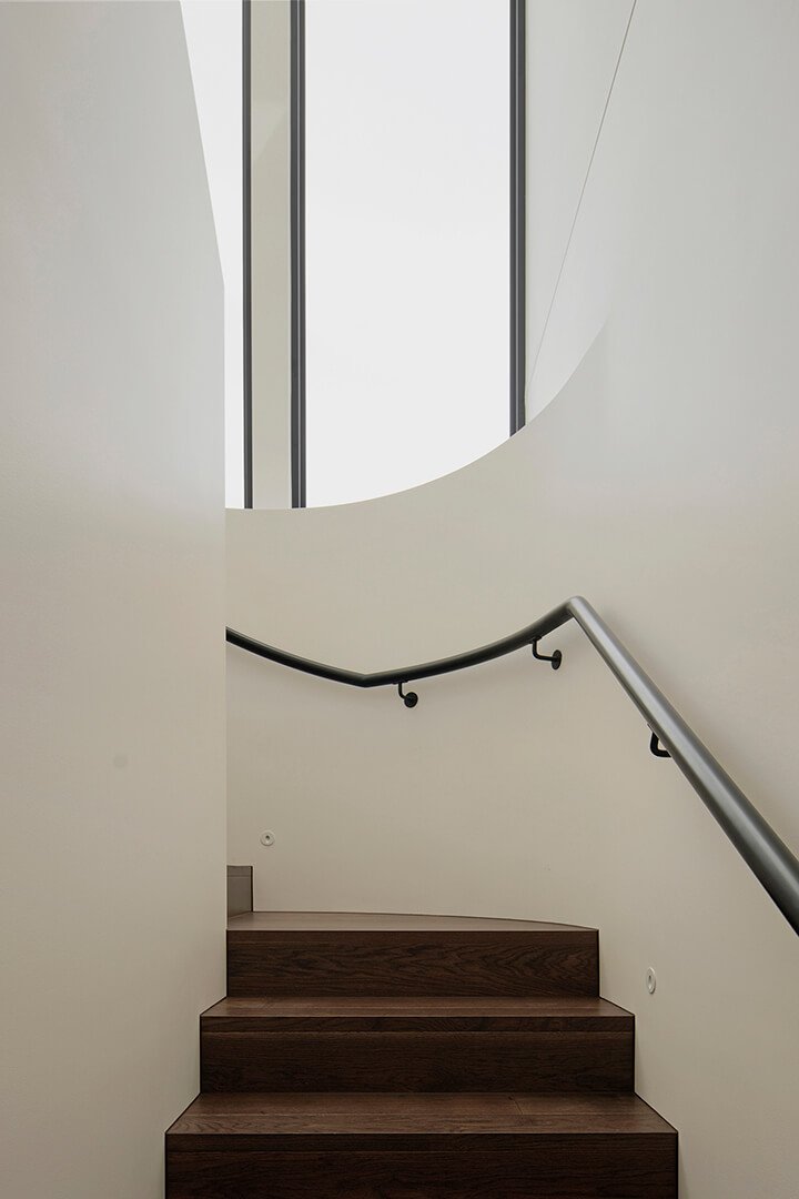Curved staircase with dark wooden steps and a sleek black metal railing. Tall vertical windows illuminate the minimalist, yet sleak design. Modern elegance.