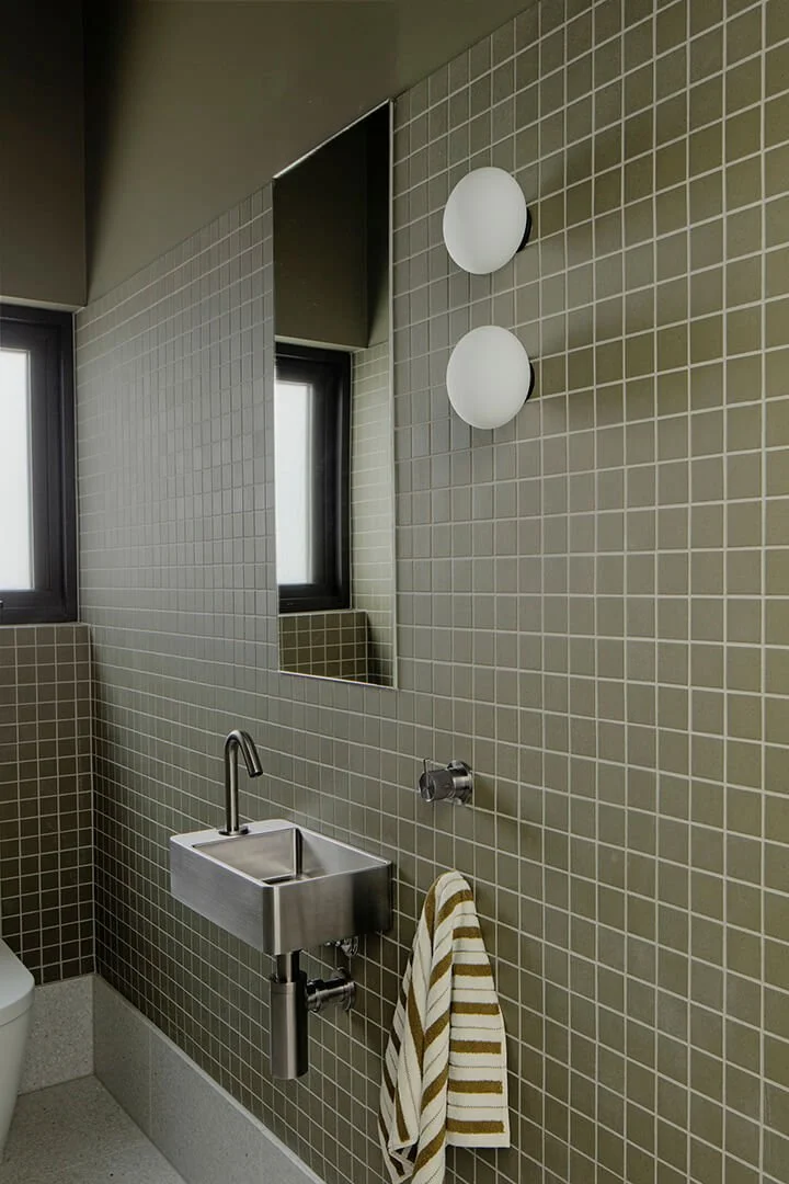 Minimalist bathroom with olive green tiles, a sleek metal sink. Two round lights beside a mirror create a modern, serene atmosphere.
