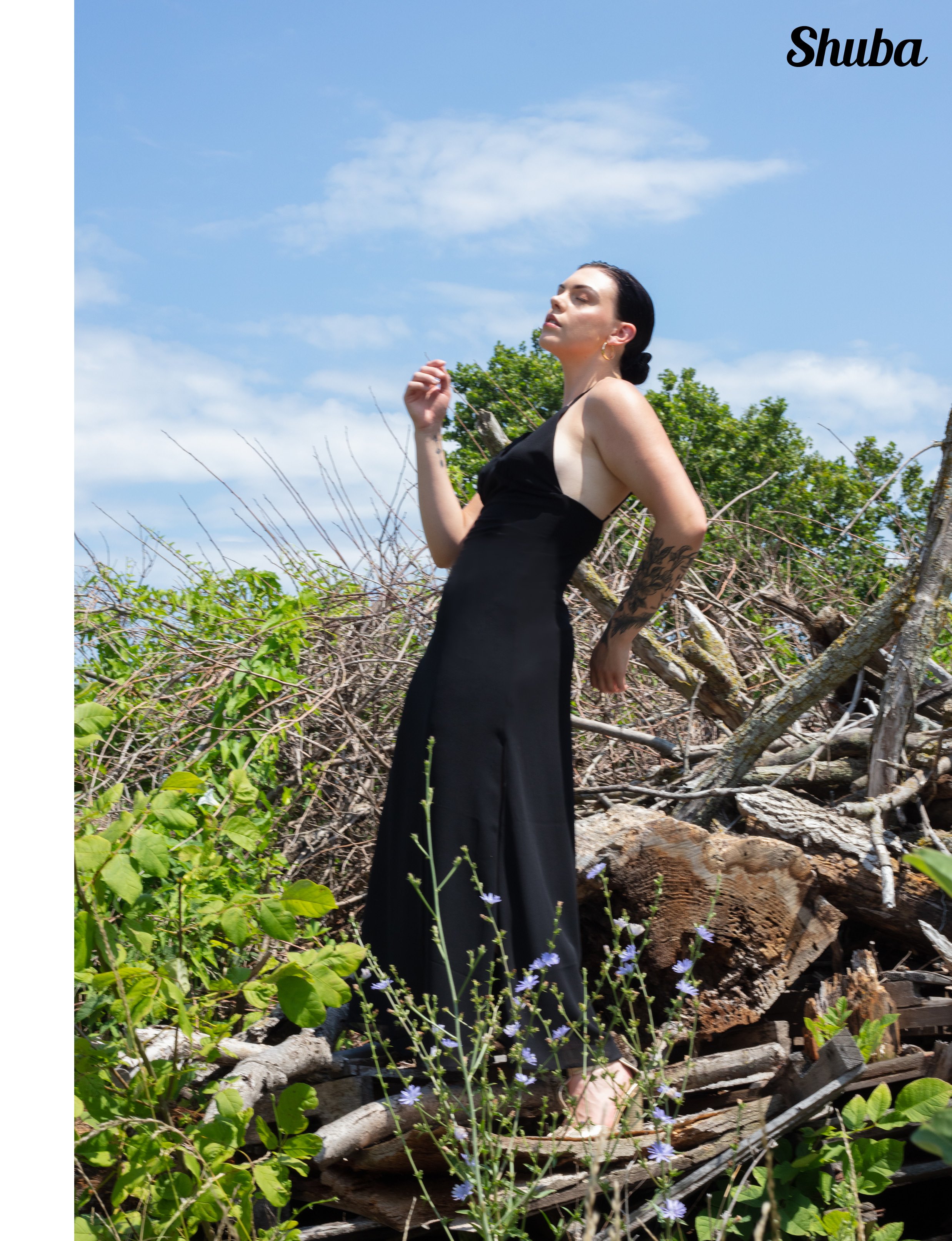 A woman in a black dress standing on a pile of logs and branches outdoors, with green foliage and purple flowers nearby, under a blue sky with scattered clouds.