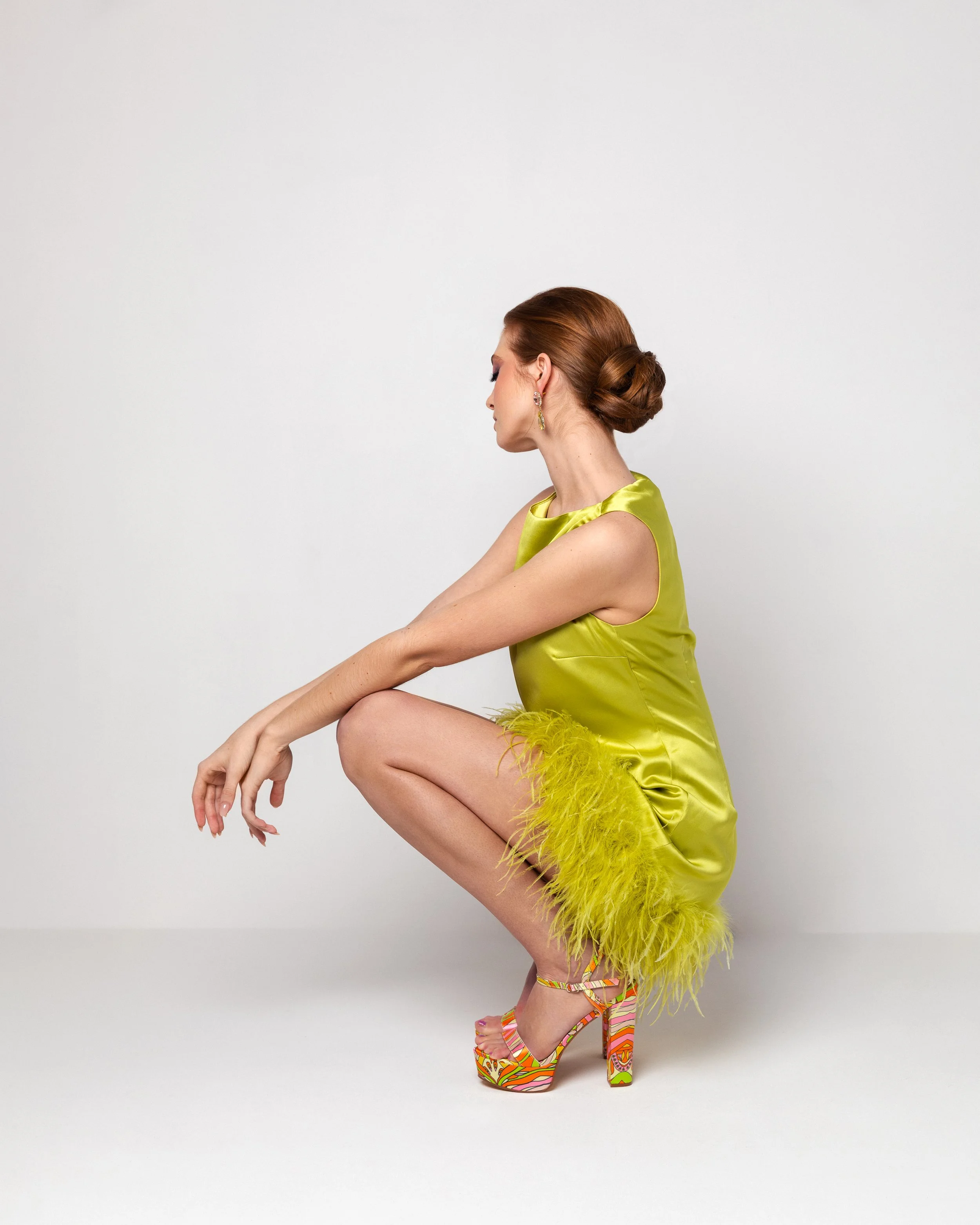 A woman with red hair styled in a bun, wearing a shiny yellow-green dress with feathered trim at the bottom, and colorful high-heeled sandals, squatting against a plain white background.