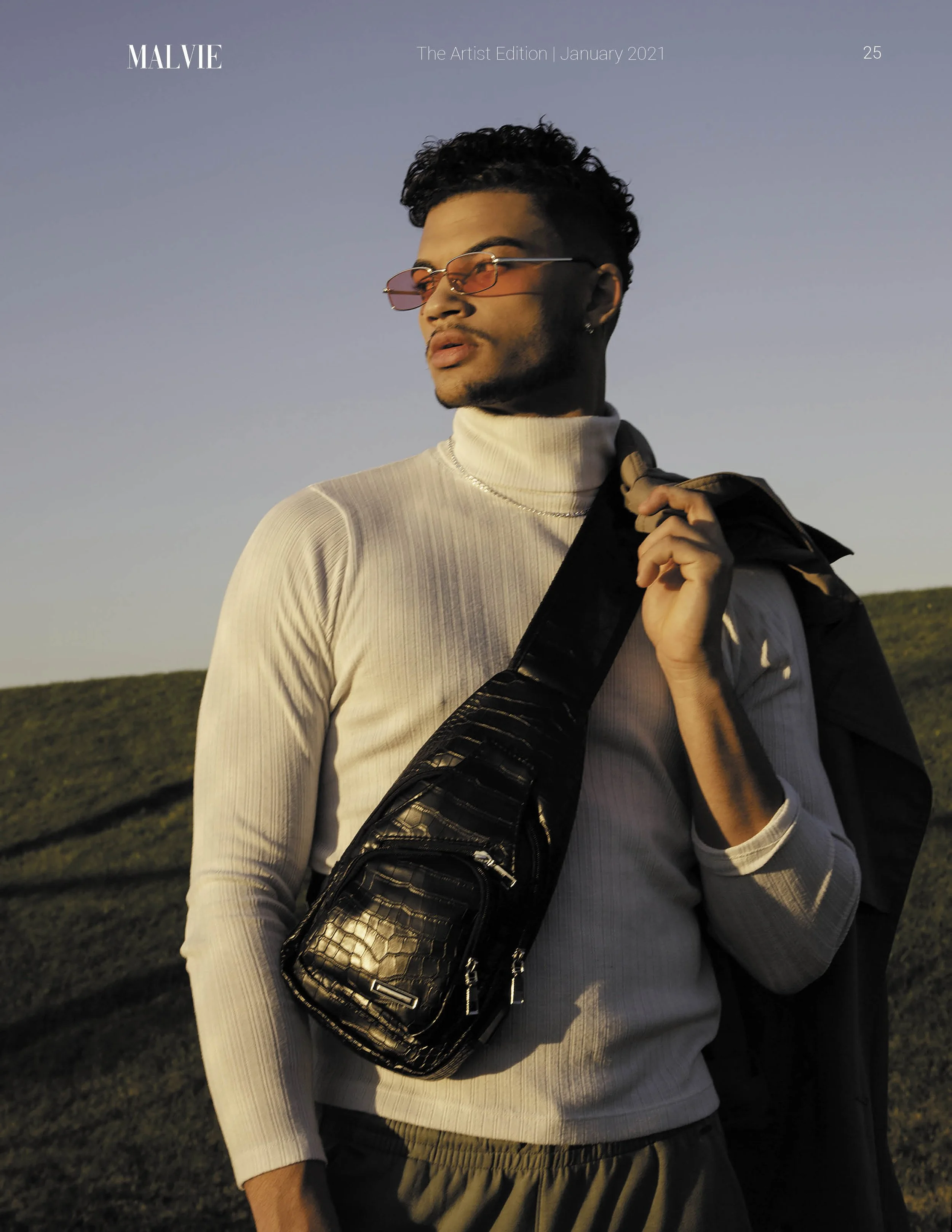 A young man stands outdoors during golden hour, wearing a white turtleneck sweater, reflective sunglasses, and a chain necklace. He has a black textured crossbody bag and a black jacket draped over his shoulder, with a grassy hillside in the backgrou
