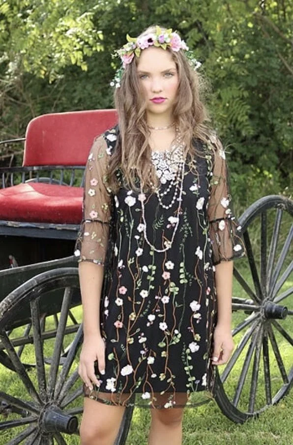 A young woman with long curly hair wearing a flower crown, a black dress with floral embroidery, and layered necklaces, standing outdoors in front of a vintage horse-drawn carriage surrounded by greenery.