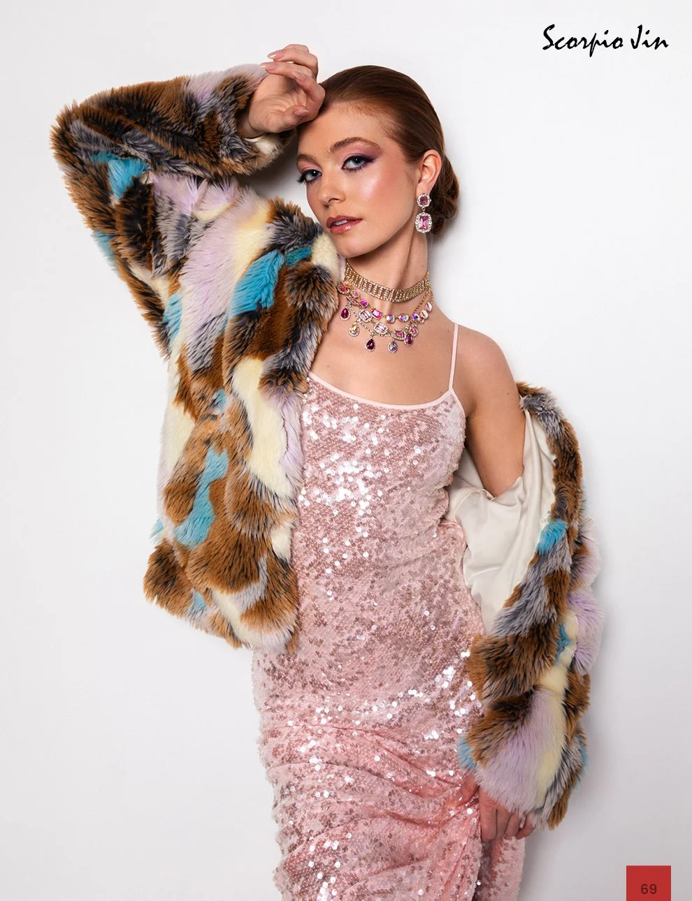 A woman posing against a white background, wearing a sequin pink dress, a colorful faux fur jacket with patches of blue, purple, yellow, and brown, and accessorized with large earrings and layered necklaces.