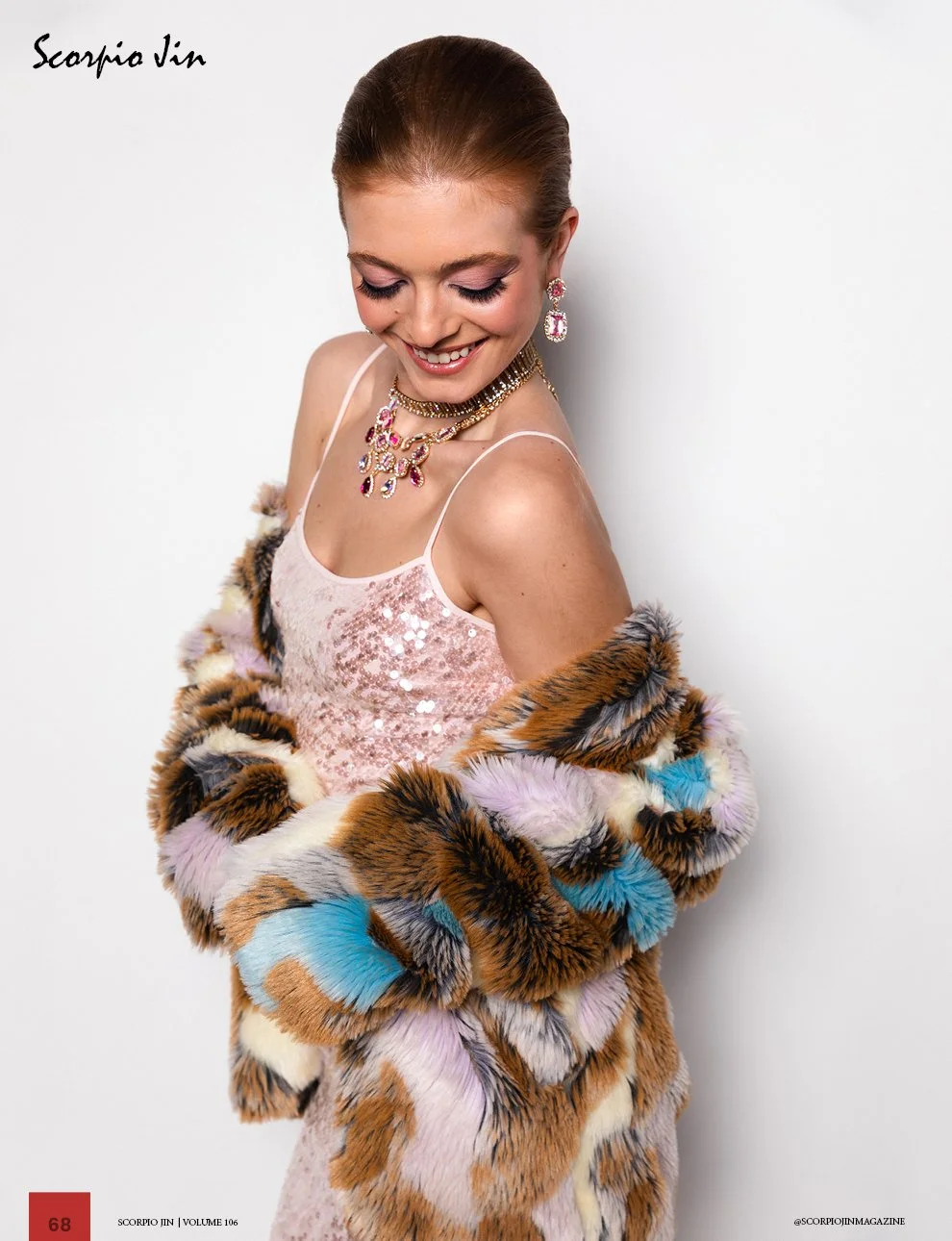 A woman smiling and looking down, wearing a pink sequin dress, a colorful fur coat with brown, pink, black, white, yellow, purple, and blue patches, and matching pink and purple jewelry, against a plain white background.