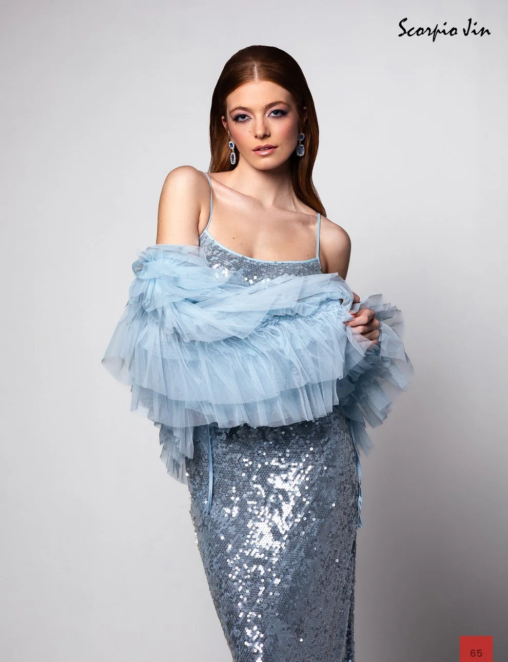 A woman with red hair wearing a silver sequin dress and earrings, holding a light blue tulle shawl, standing against a plain background with "Scorpio Jin" written in black script in the top right corner.
