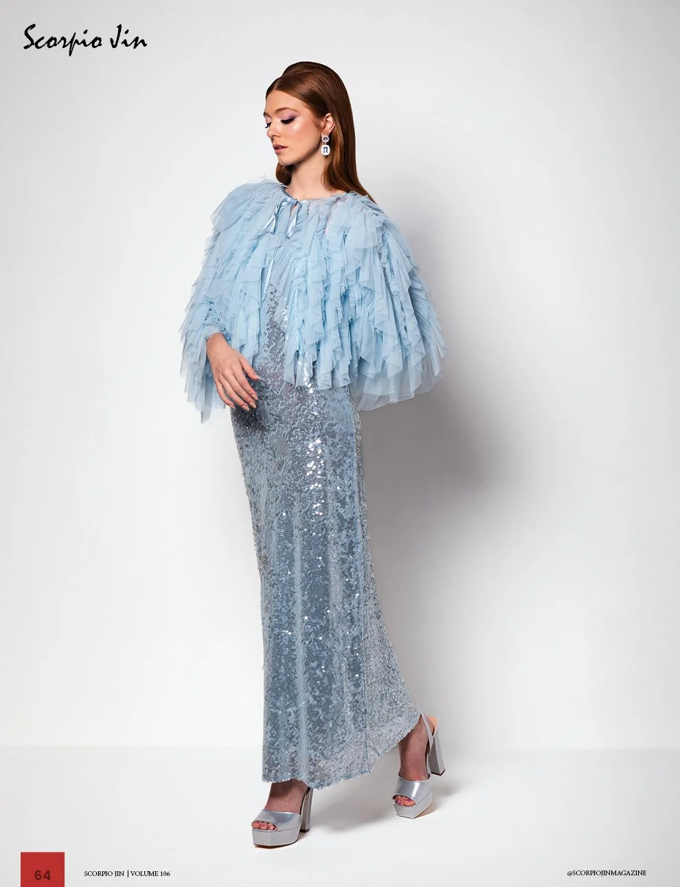 Woman modeling a haute couture dress with a sparkly silver sequined skirt and a light blue ruffled cape-like top, wearing silver platform heels and statement earrings, standing against a plain white background.