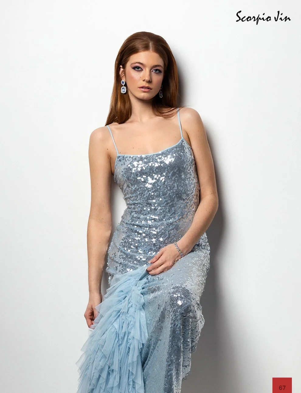 A young woman with long red hair wearing a silver sequined dress with spaghetti straps, earrings, and a bracelet, standing against a white wall, holding a light blue tulle fabric. The top right corner has the signature 'Scorpio Jin'.