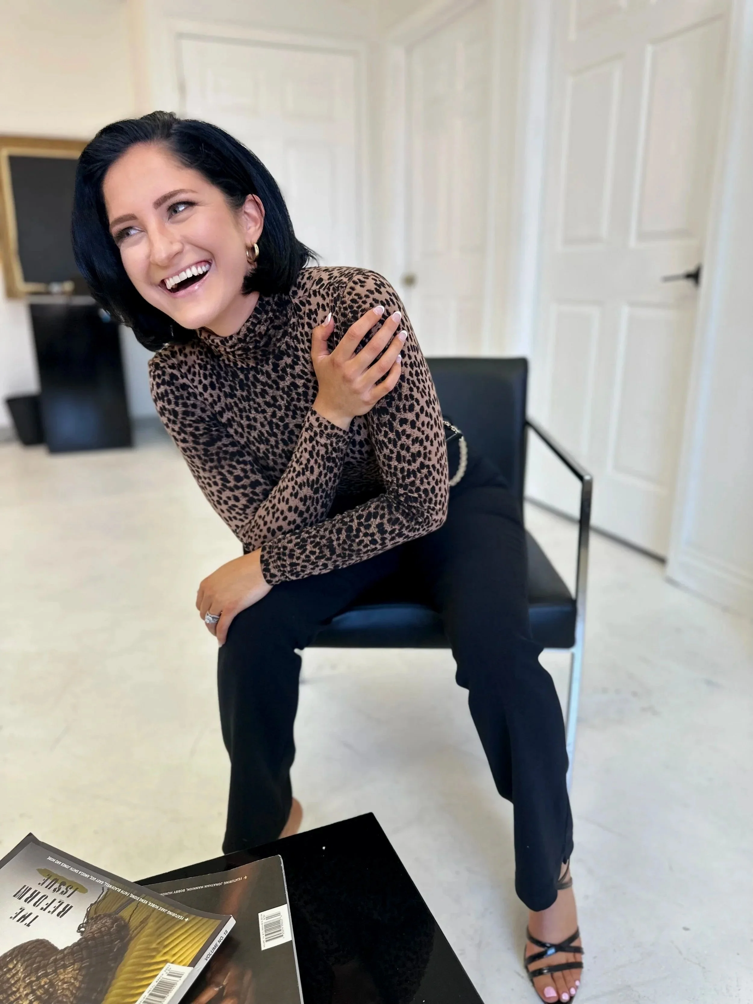 A woman with black hair smiling while sitting on a black chair in a bright room, wearing a leopard print top and black pants.