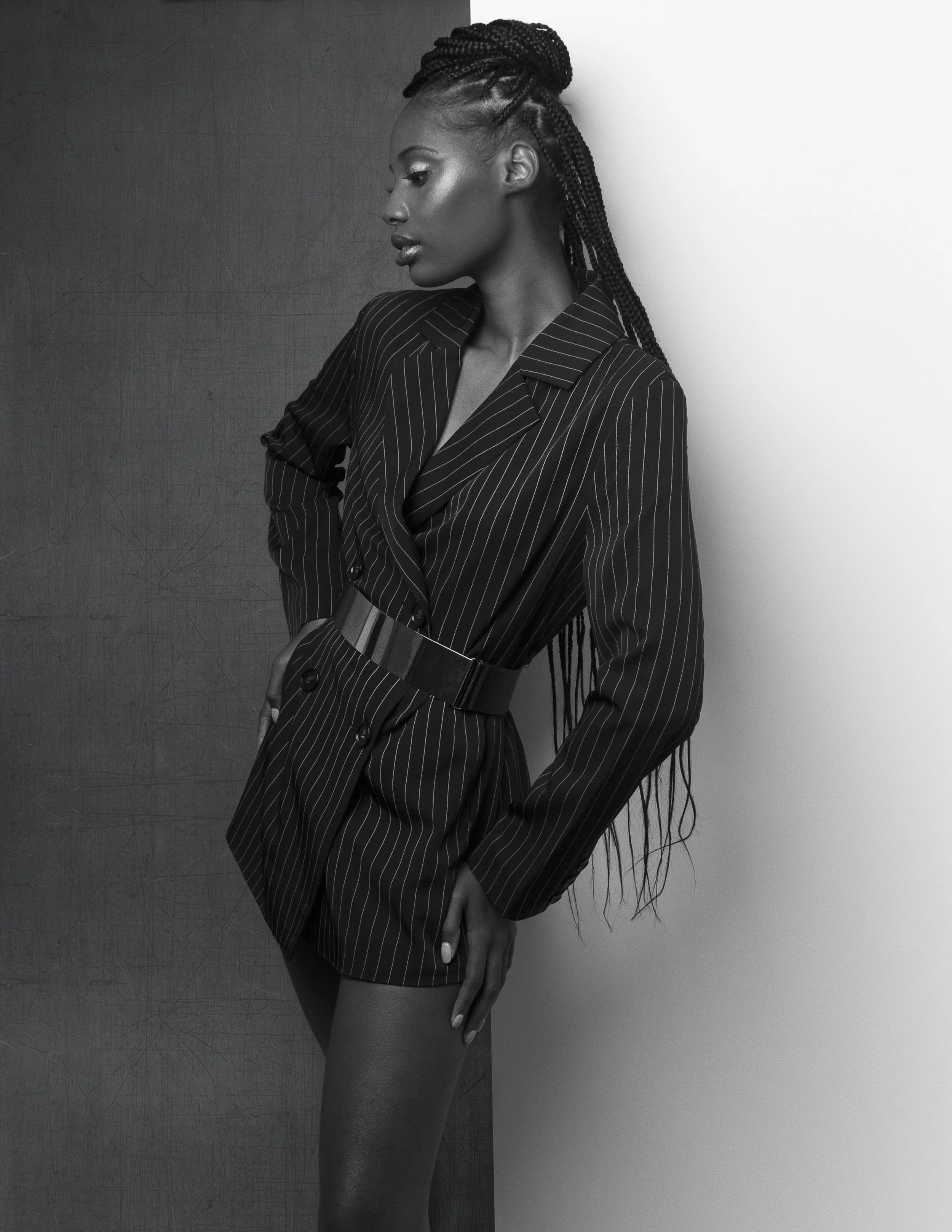 Black and white photo of a woman with braided hair wearing a pinstripe blazer dress with a belt, posing against a contrasting background.