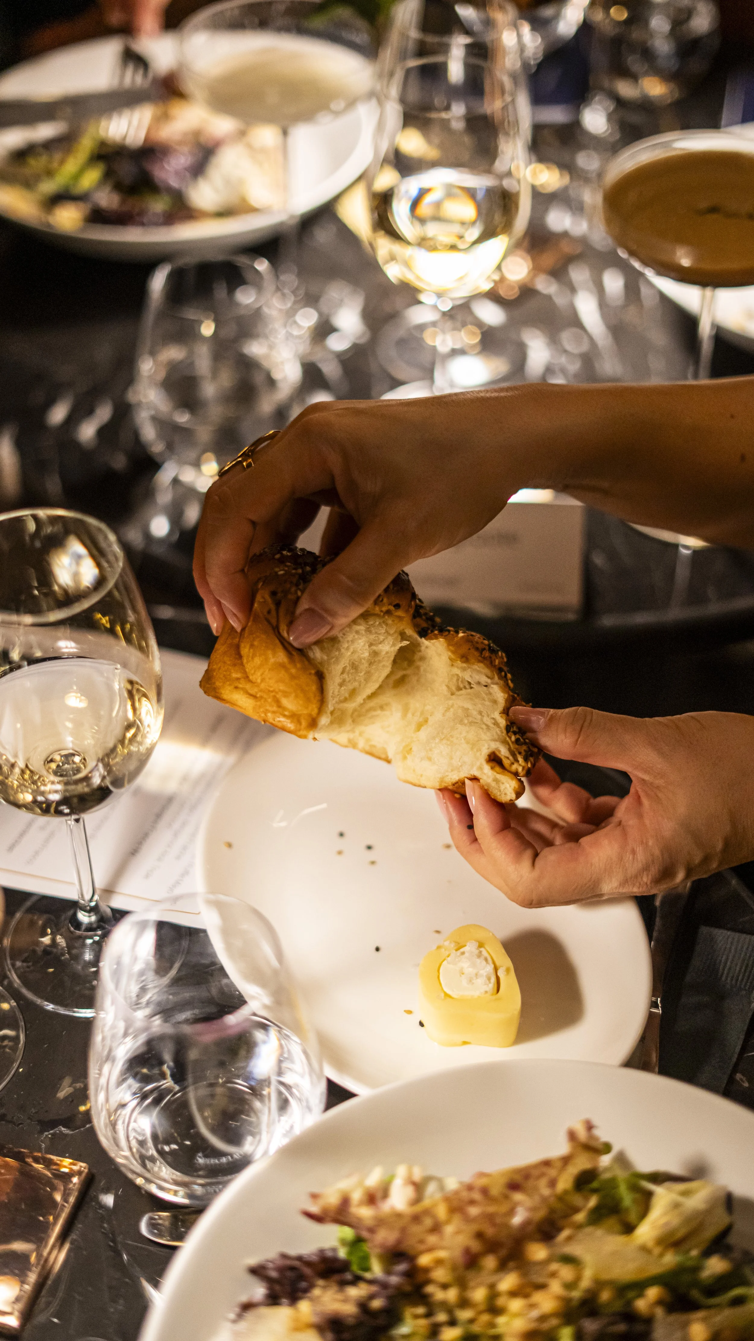 Someone ripping a piece of bread with the background of lots of food at wine glasses.