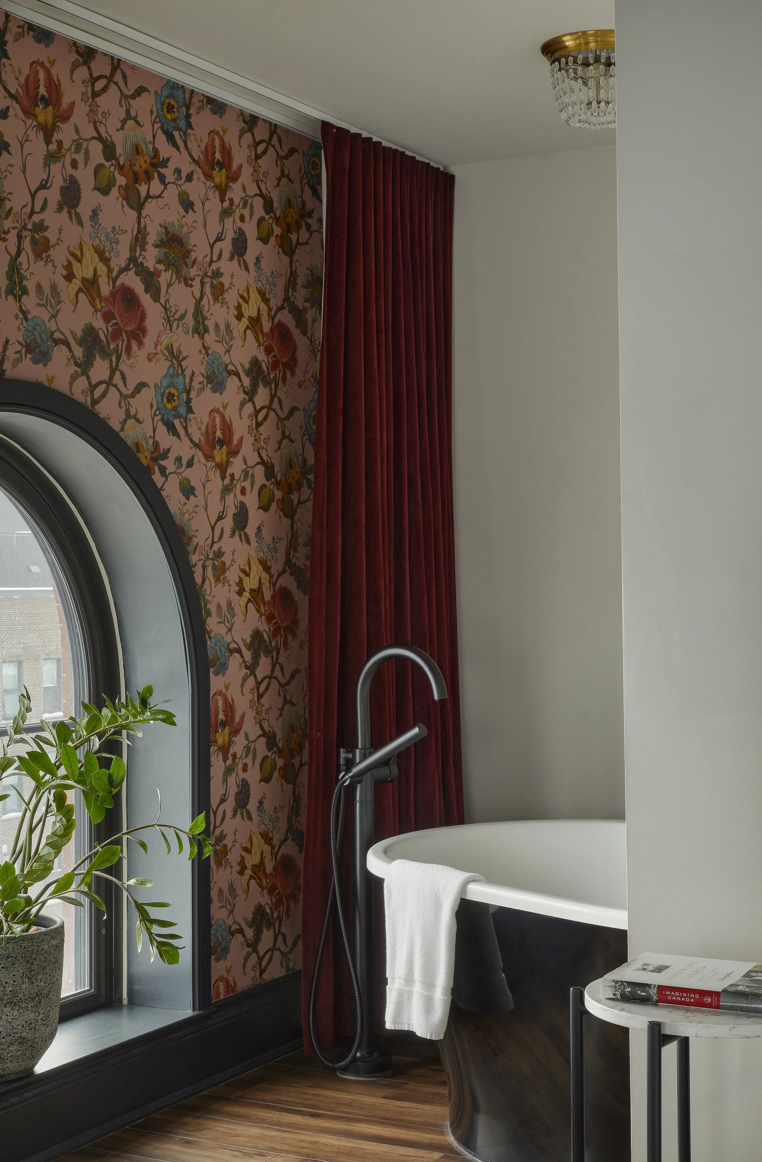 The corner of a bathroom with an edgy pink floral wall paper and a dark claw foot tub.