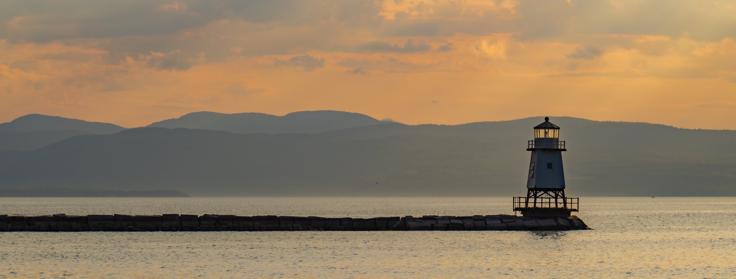 A lighthouse on a breakwater at sunset with mountains in the background and a partly cloudy sky.