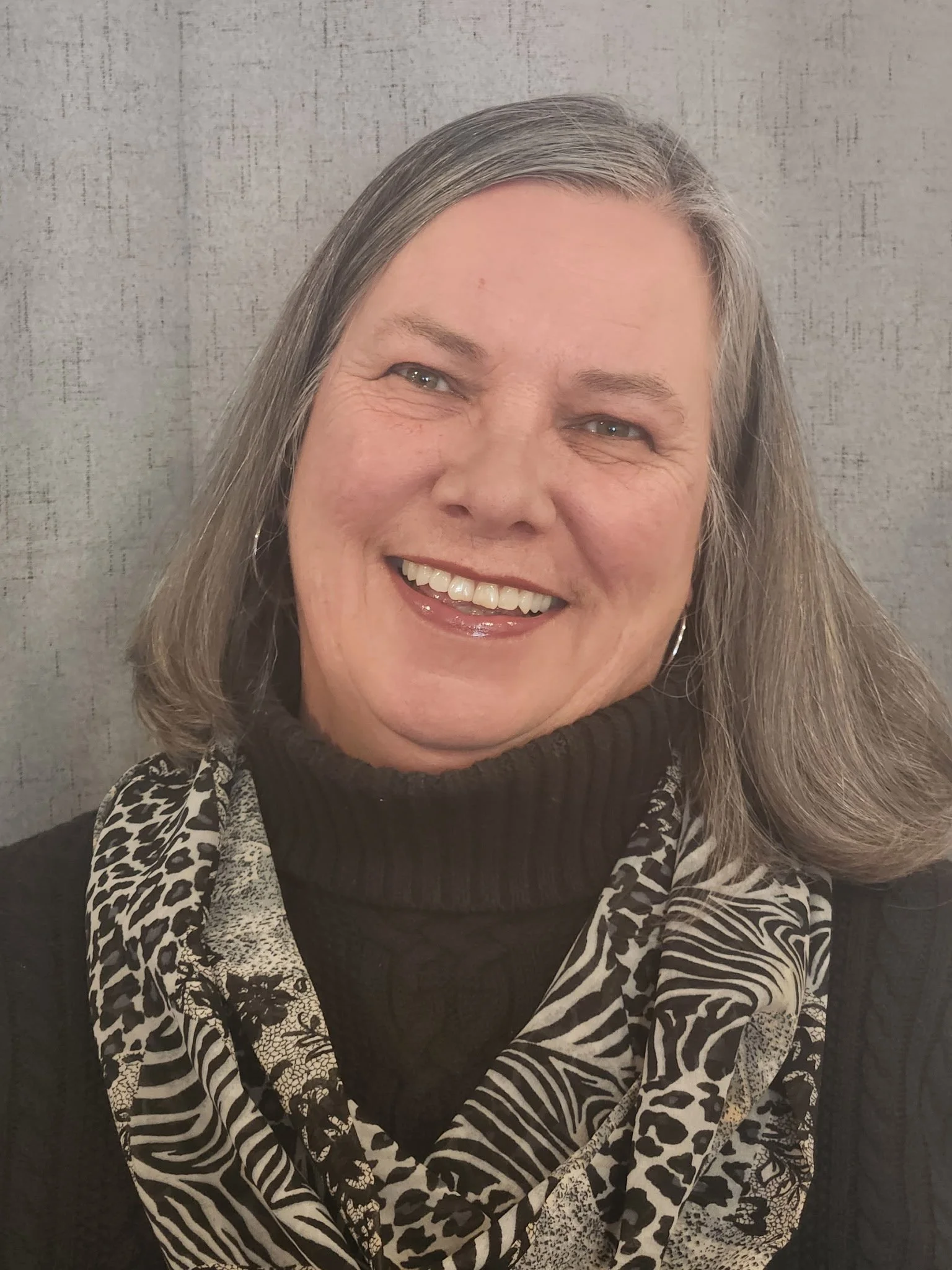 A middle-aged woman with shoulder-length gray hair, smiling, wearing a black turtleneck sweater and a patterned scarf with animal print.