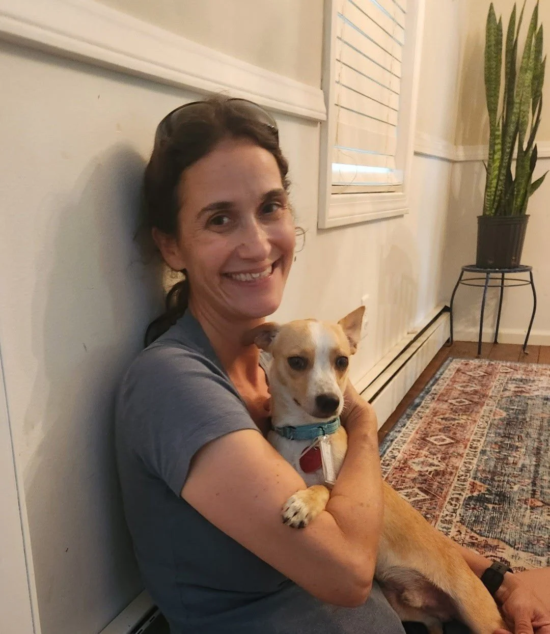 A woman sitting on the floor, smiling, and holding a small tan and white dog in her arms inside a house.