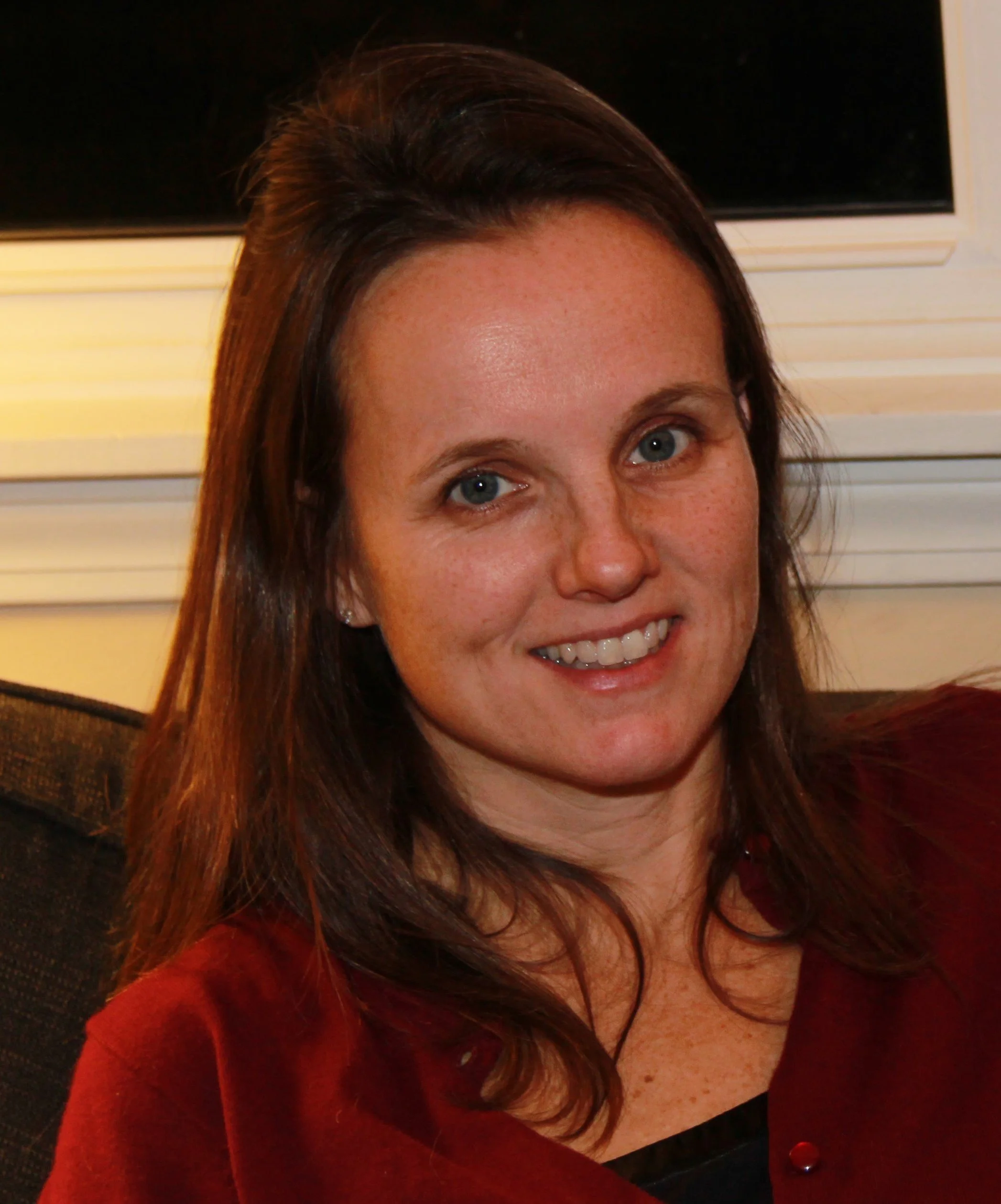 A woman with light skin, brown hair, and blue eyes smiling, sitting indoors in front of a TV and window.