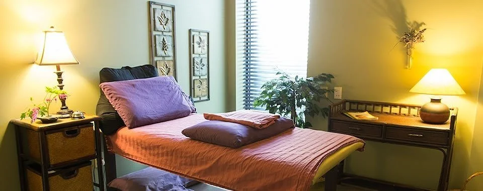 A cozy bedroom with a bed covered in pink and purple bedding, two lamps on side tables, wall art, and a window with blinds.