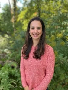 A woman with long dark hair smiling, wearing a pink sweater outdoors in a green, wooded area.