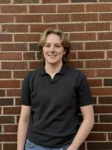 A young woman with short blonde hair standing in front of a brick wall, smiling, wearing a black polo shirt and blue jeans.