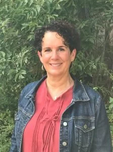 A woman with short curly hair smiling outdoors, wearing a pink top and denim jacket, standing in front of green foliage.