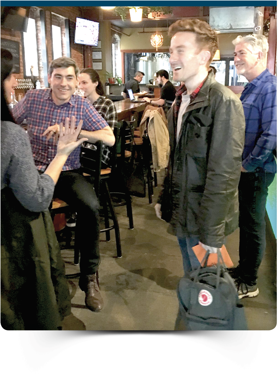 Group of people socializing in a bar, with one person holding a backpack and others seated at the bar counter.
