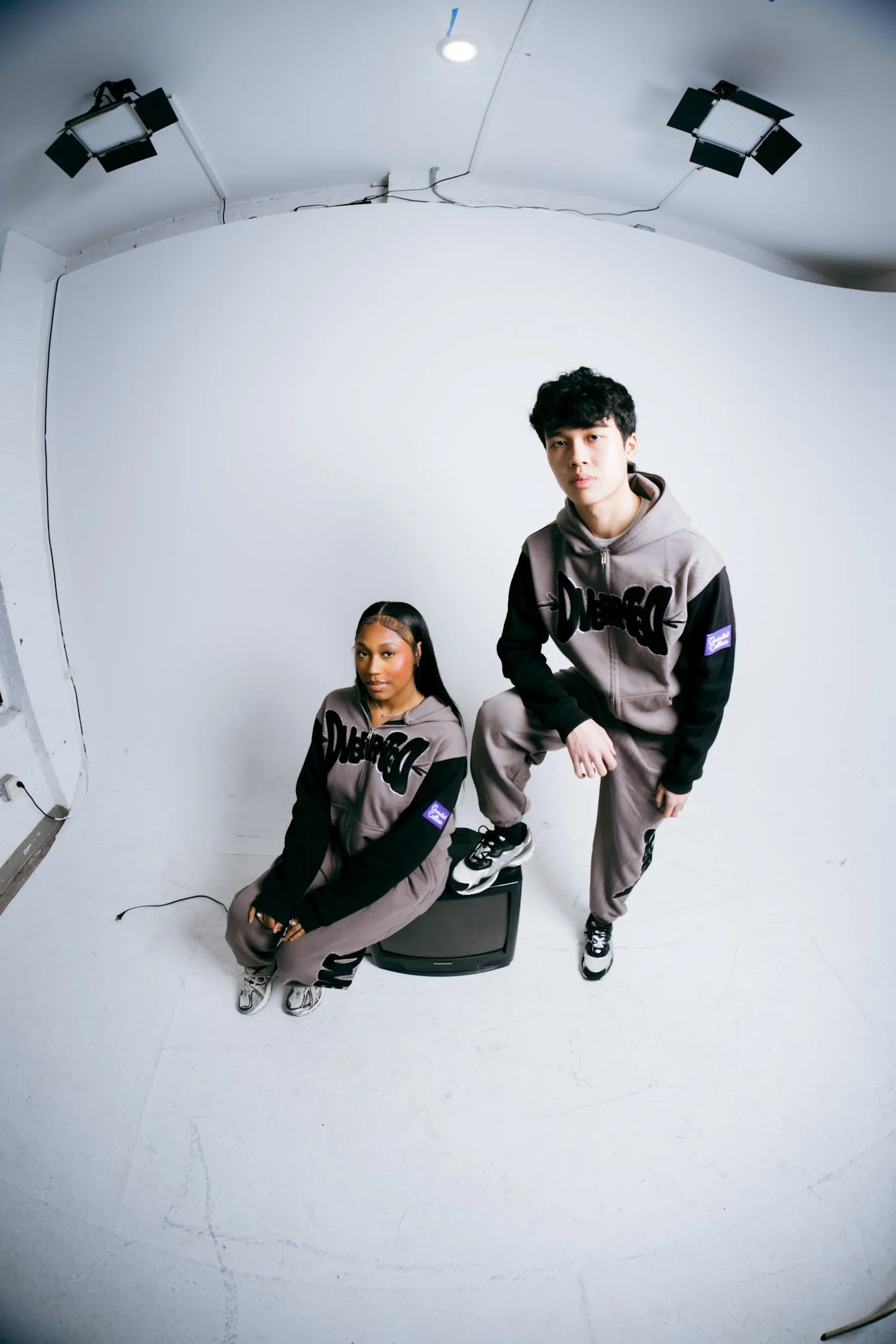 Two young individuals wearing matching gray and black hoodies and gray pants in a photography studio with a white background. One is seated on a small television set, and the other is standing with one foot on the TV. The studio has studio lights mounted on the ceiling.