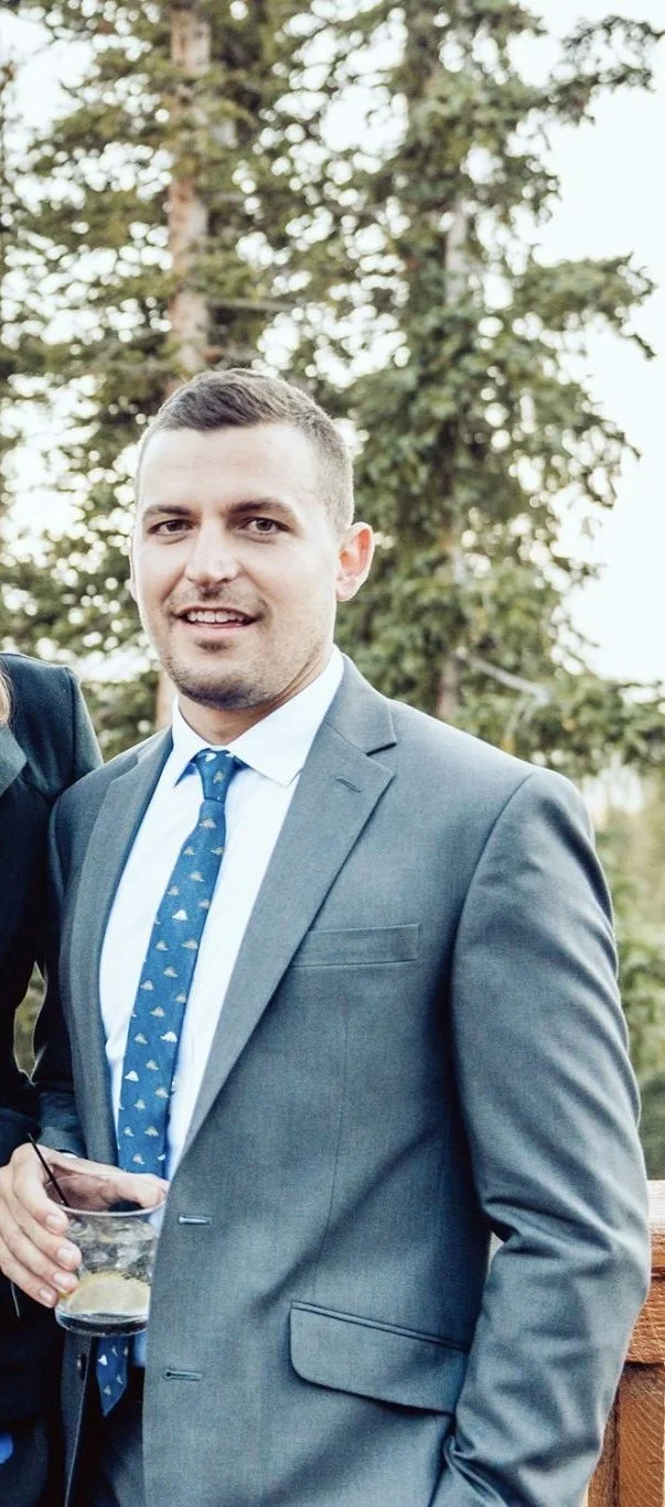 A man in a blue suit and tie standing outdoors in front of tall trees, smiling at the camera.