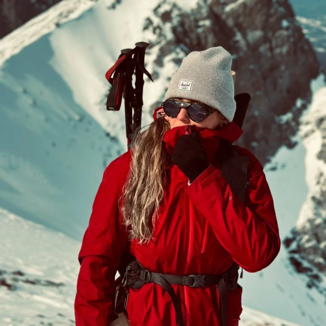 A woman dressed in red winter gear, wearing a gray beanie and black sunglasses, standing in a snowy mountain landscape with a backpack and an ice axe on her back.