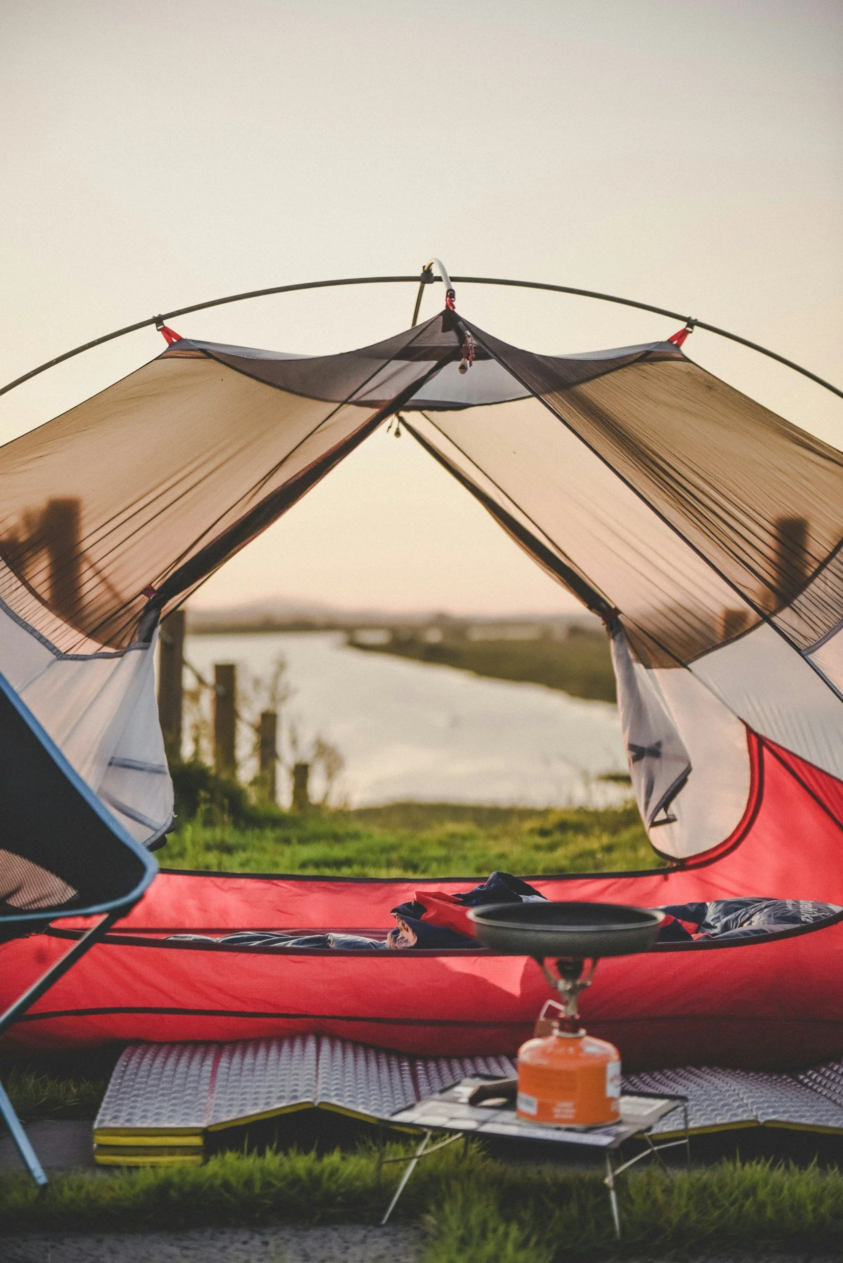 View from inside a tent set up near a body of water during sunset, with camping gear and a portable stove in the foreground.
