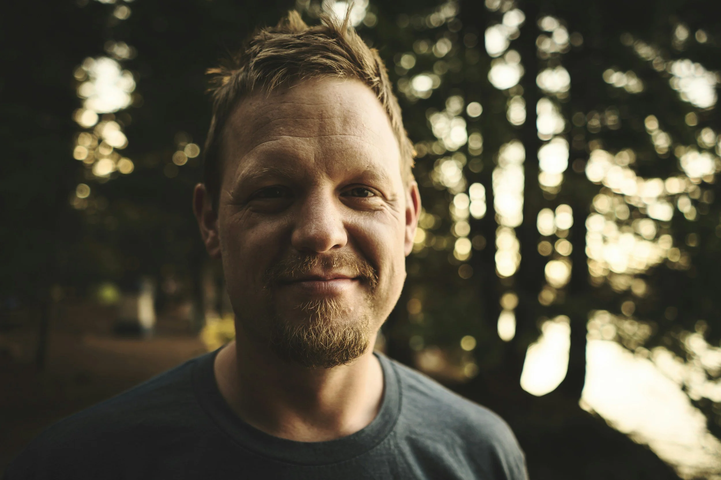 Close-up of a smiling man with light brown hair and a beard, standing outdoors during sunset with blurred trees and warm light in the background.