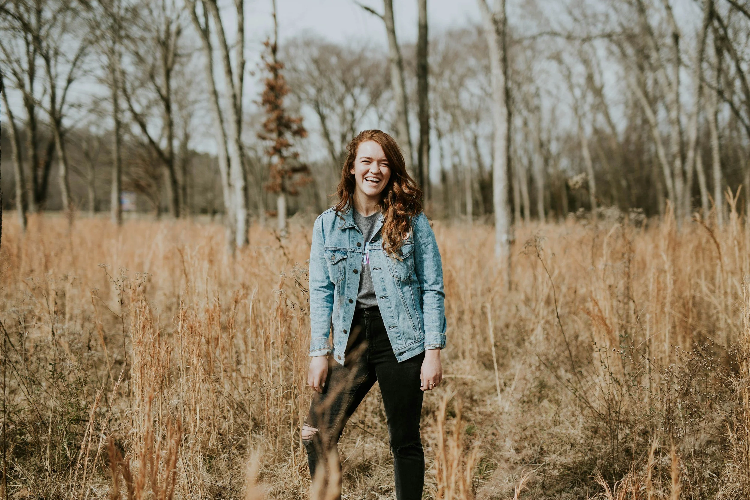 A young woman with long brown hair, wearing a denim jacket and ripped black jeans, standing in a field of tall, dry grass with trees in the background, smiling and laughing.