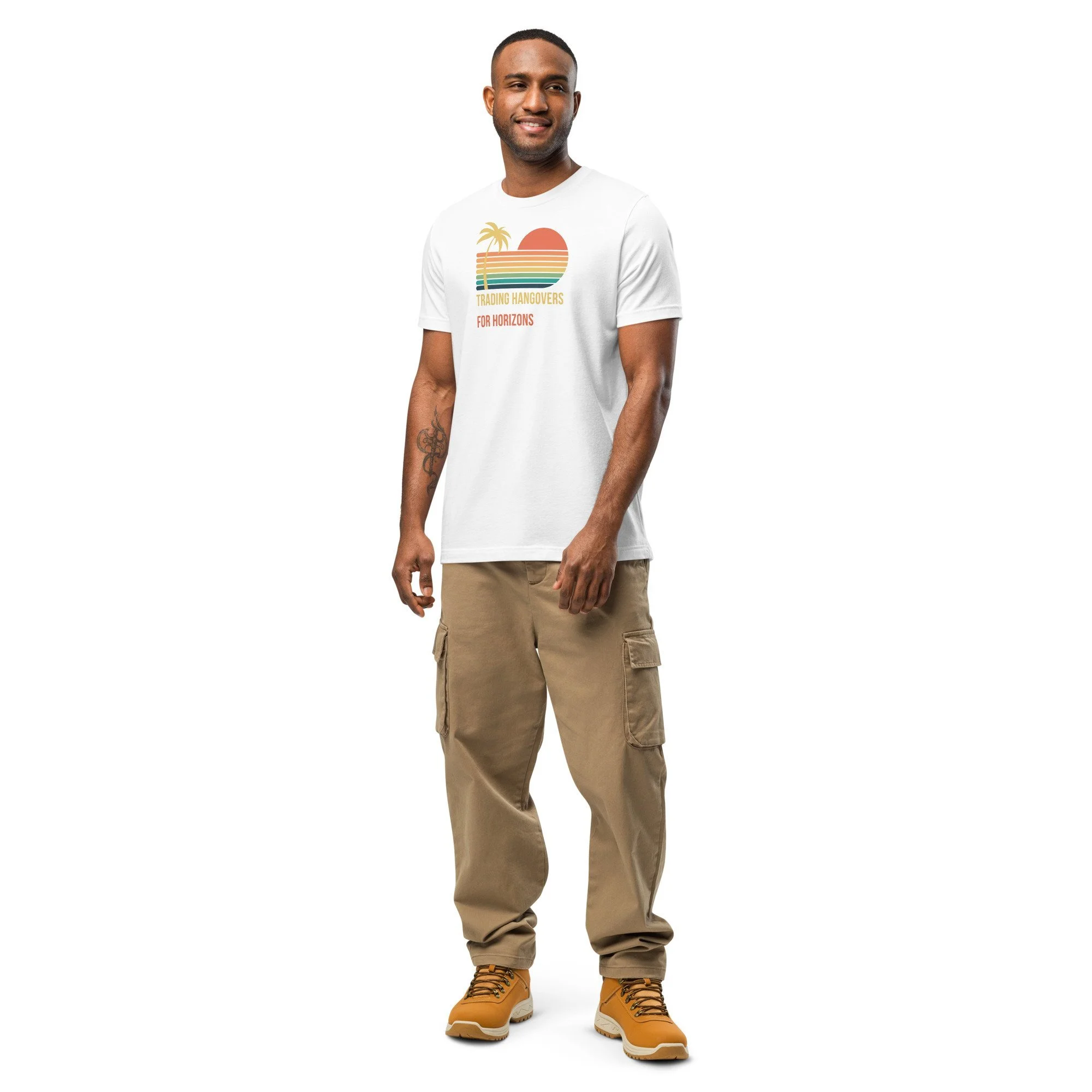 A young man with short hair and a tattoo on his right forearm, wearing a white T-shirt with a colorful sunset and palm tree design, tan cargo pants, and tan hiking boots, standing against a plain white background and smiling.