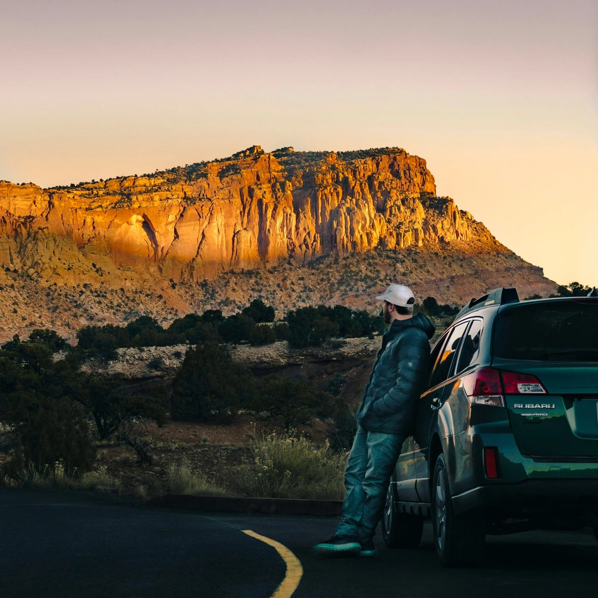 A man in a jacket and hat leaning against a dark green Subaru SUV parked on the side of a desert road at sunset, with a large rocky cliff in the background.