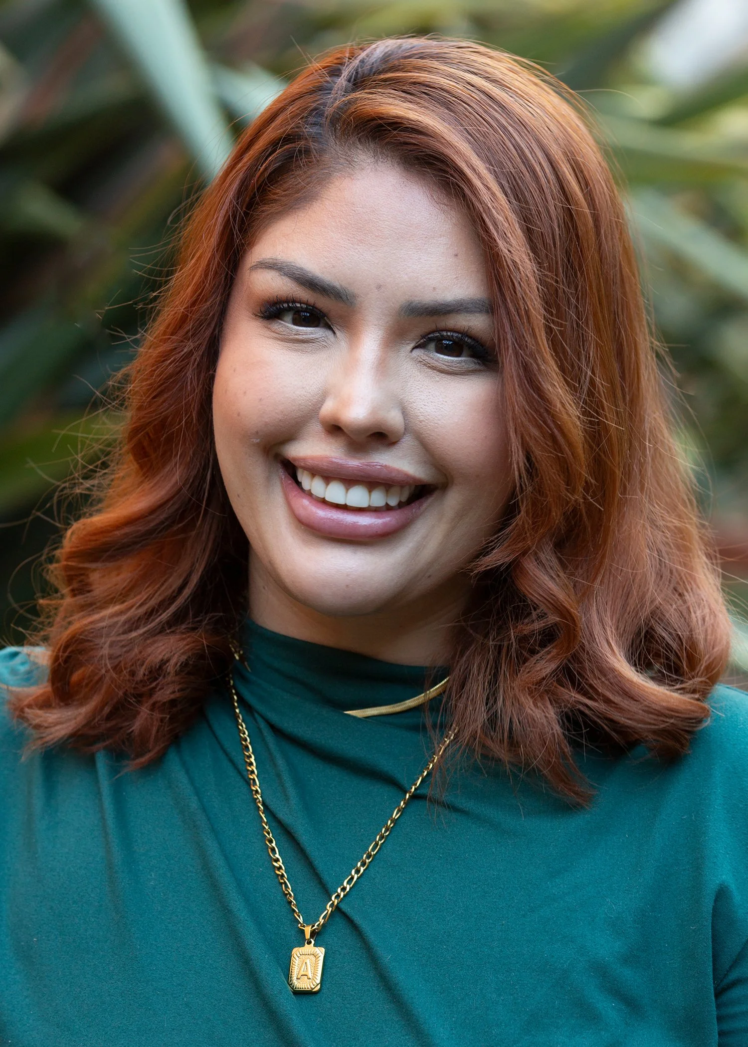 Smiling woman with long, wavy brown hair with blonde highlights, wearing a colorful patterned top, standing outdoors with green plants in the background.