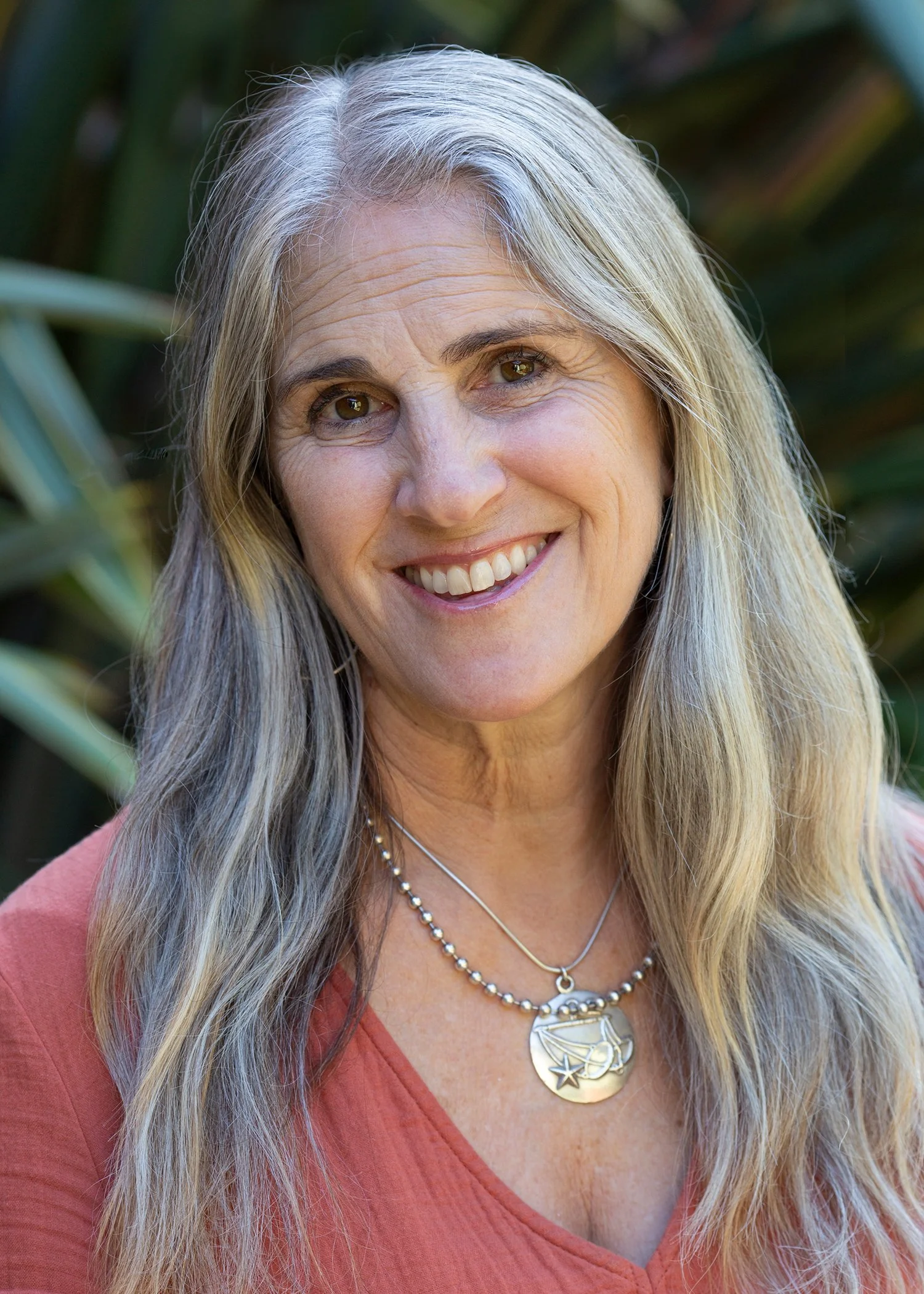 A smiling woman with long gray hair, wearing layered necklaces, hoop earrings, a green top, and a light cardigan, outdoors with plants in the background.