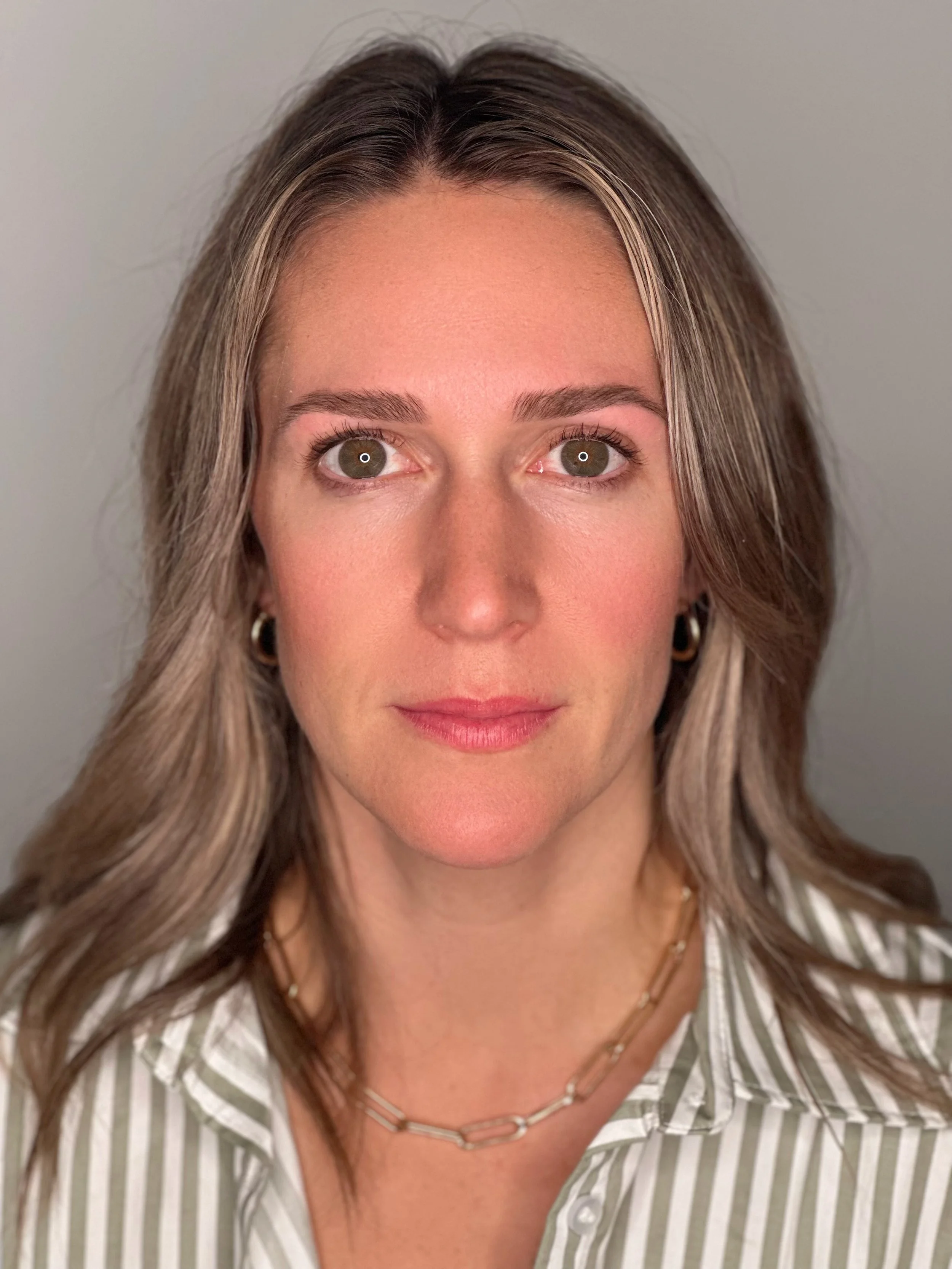 A woman with shoulder-length wavy hair, wearing a striped shirt, small hoop earrings, and a chain necklace, looking directly at the camera with a neutral expression.