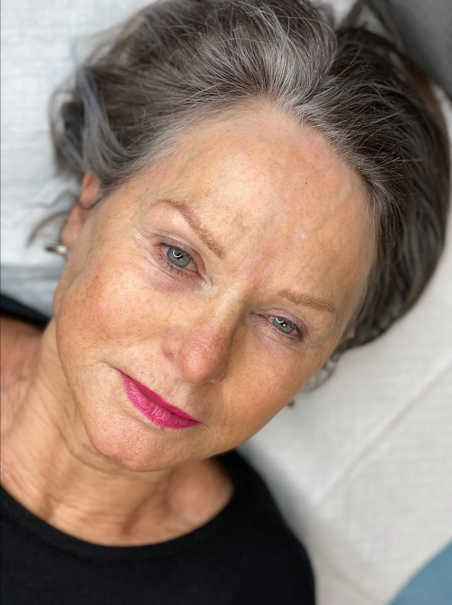 Close-up of an older woman with gray hair, light skin, freckles, wearing pink lipstick, lying down on a white pillow, looking softly at the camera.