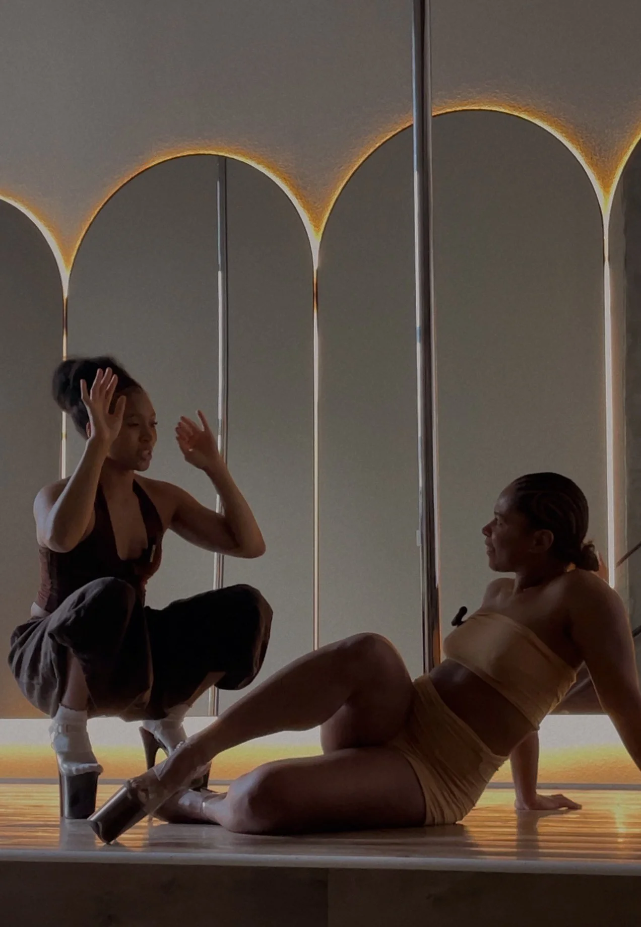 PoleBait founder Naya Somanya kneeling beside a student in platform heels, guiding her through movement during a private pole coaching session at PoleBait Haus in Los Angeles
