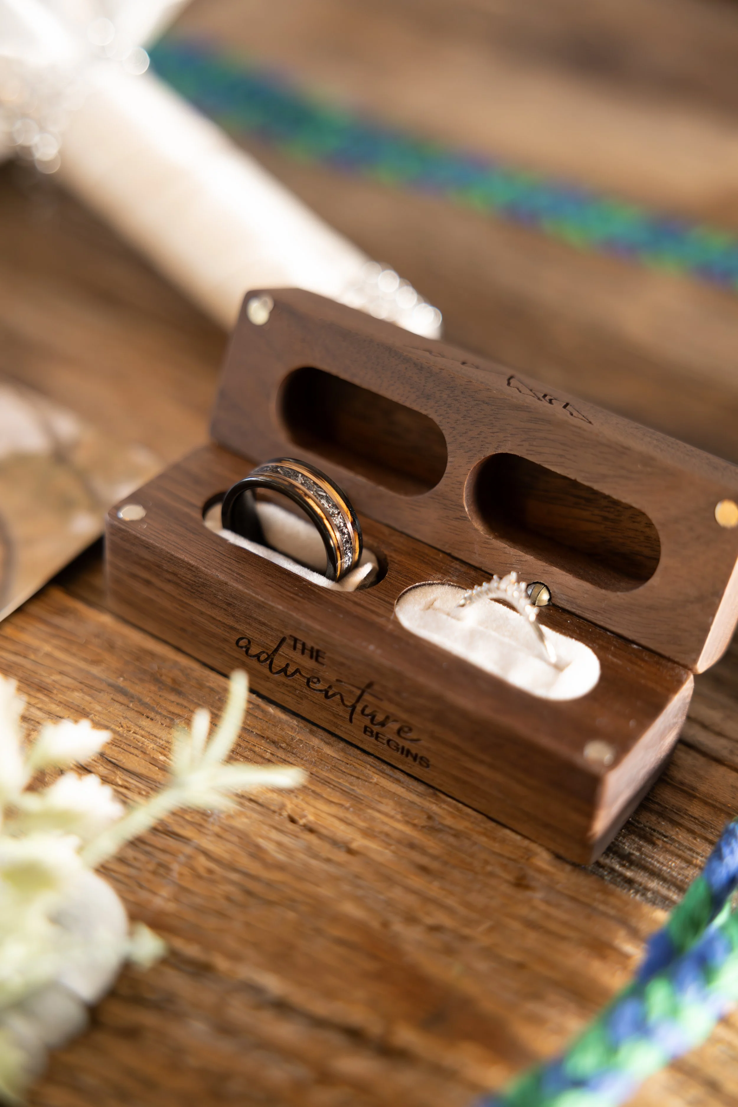 Wooden wedding ring box with engraved words 'The adventure begins', holding two rings, on a wooden surface, with a white wedding shoe and decorative elements in the background.