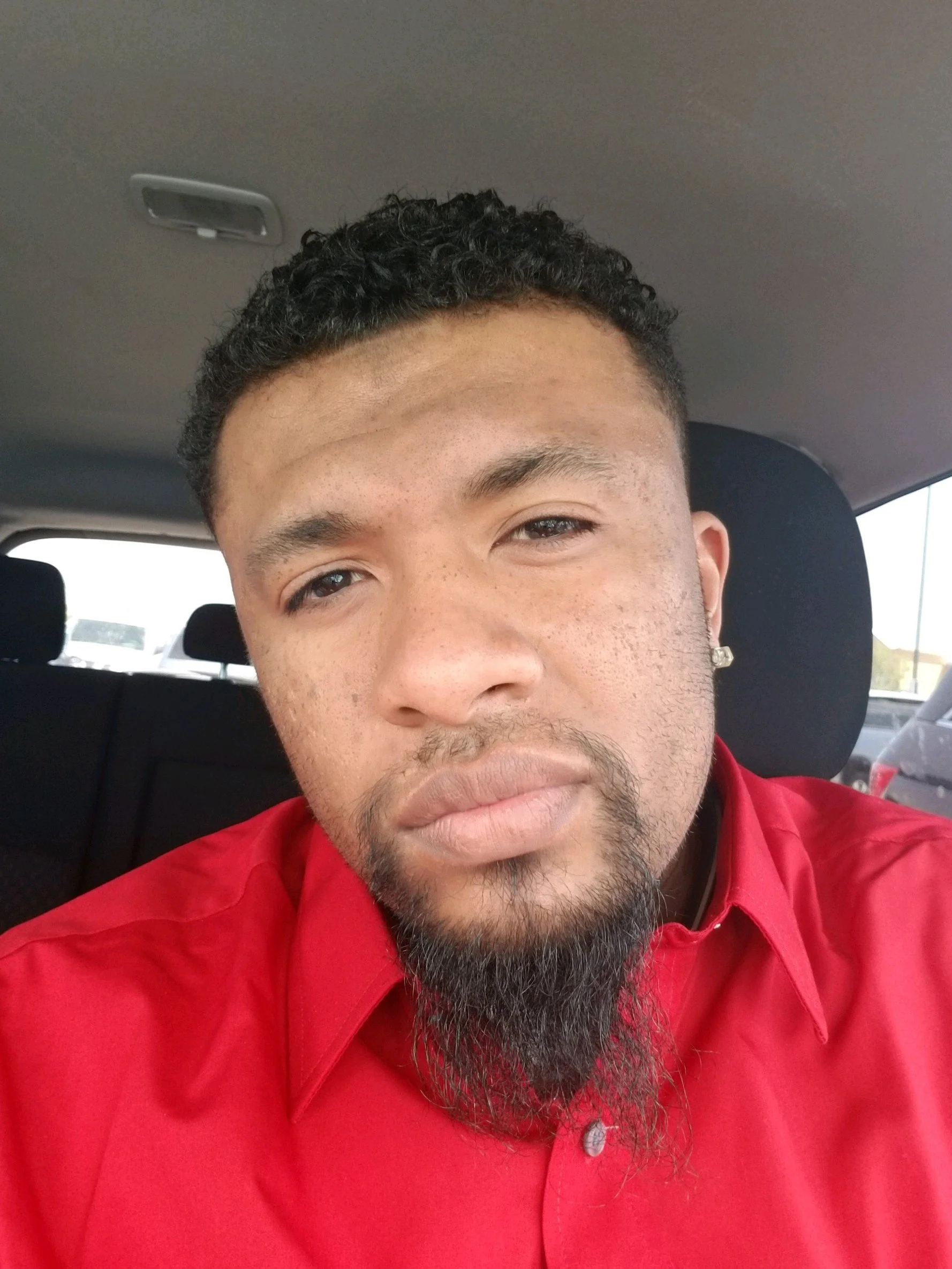 Close-up selfie of a man with short curly hair, a goatee, and diamond stud earrings, wearing a red button-up shirt, sitting inside a car with a parking lot visible in the background.