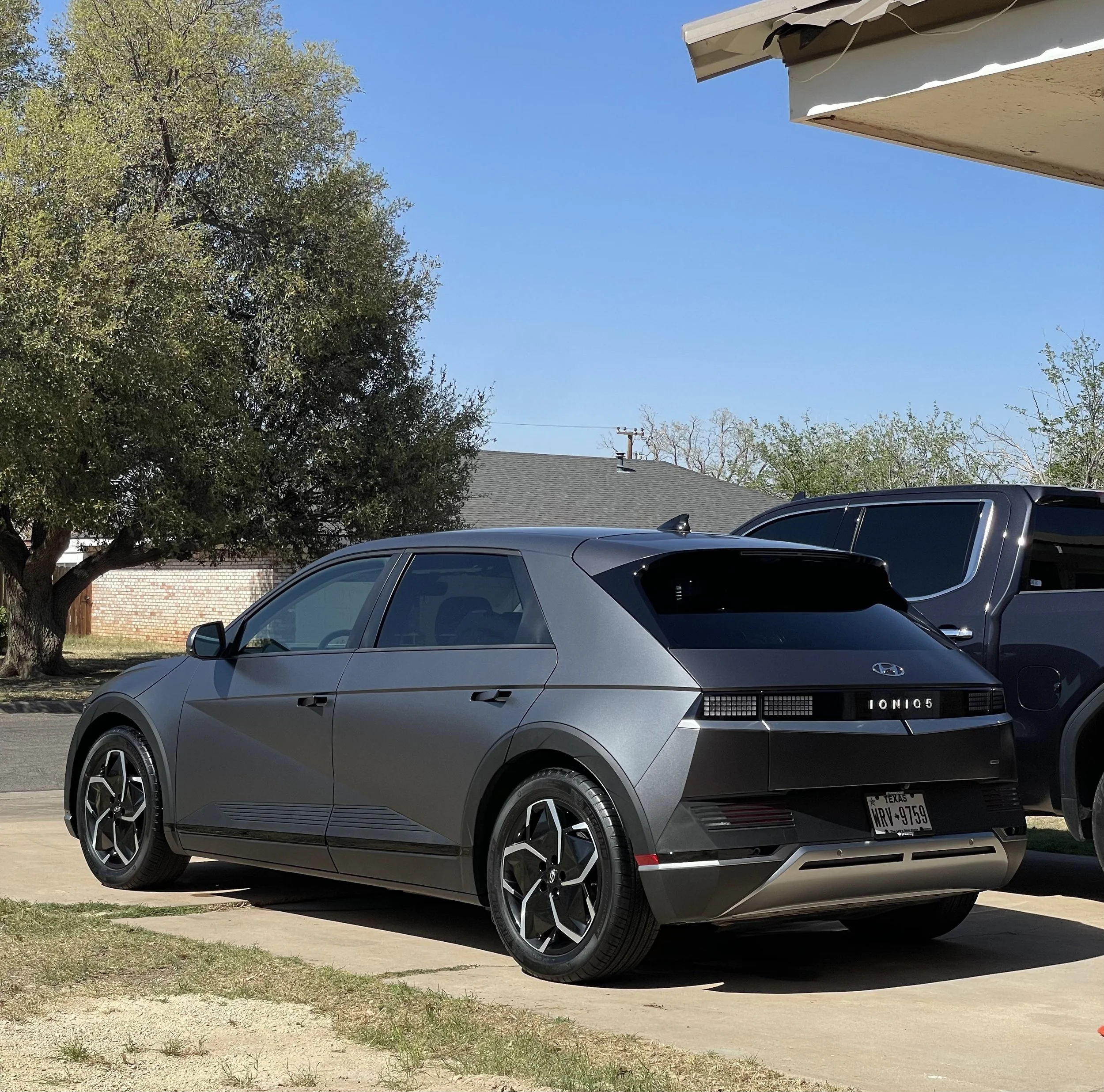 Silver Hyundai Ioniq 5 parked in front of a house with trees and a blue sky visible in the background.