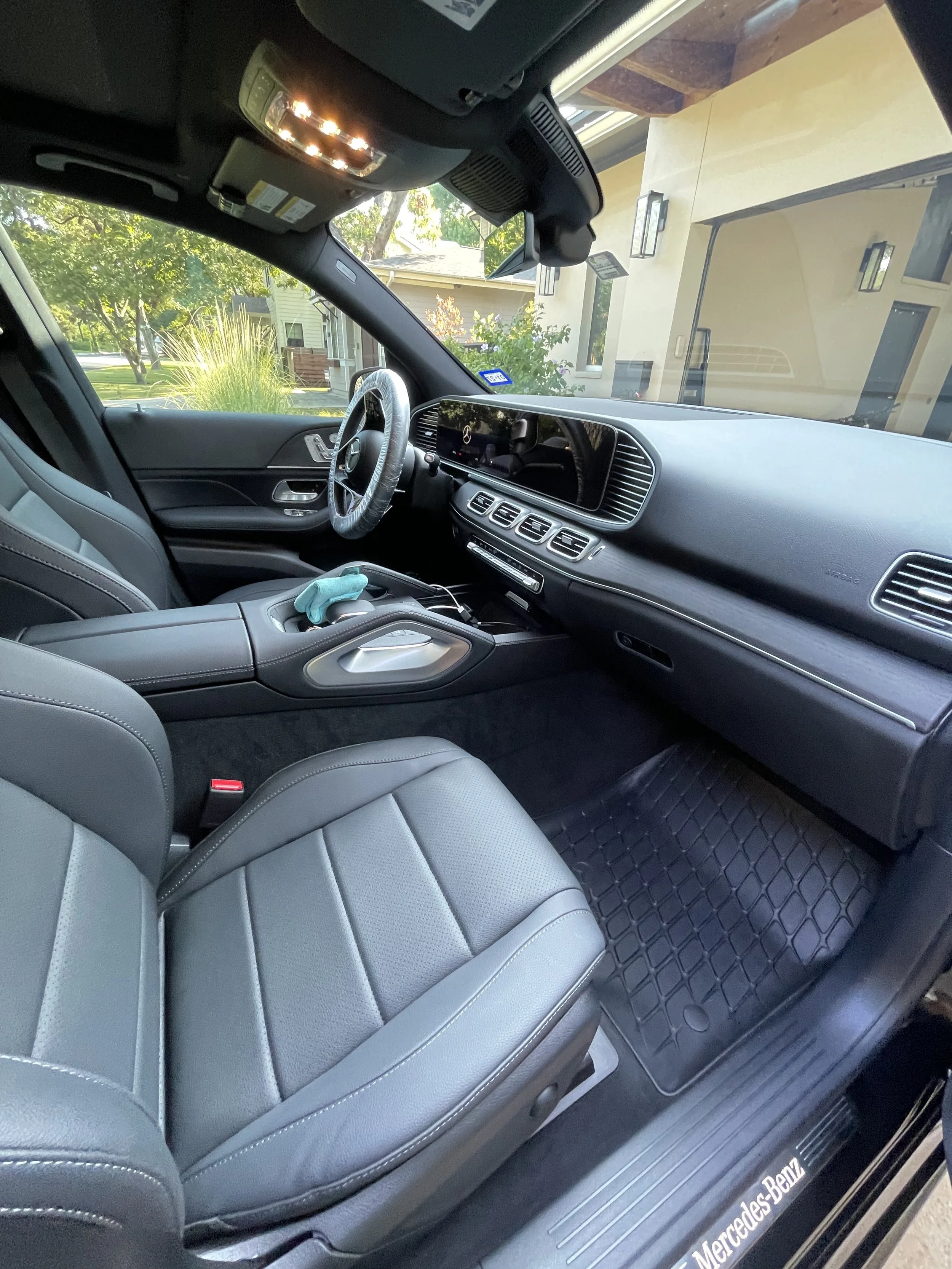 The interior of a Mercedes-Benz vehicle showing gray leather seats, a dashboard, and a steering wheel covered with plastic. The window reveals a residential neighborhood with trees and houses outside.