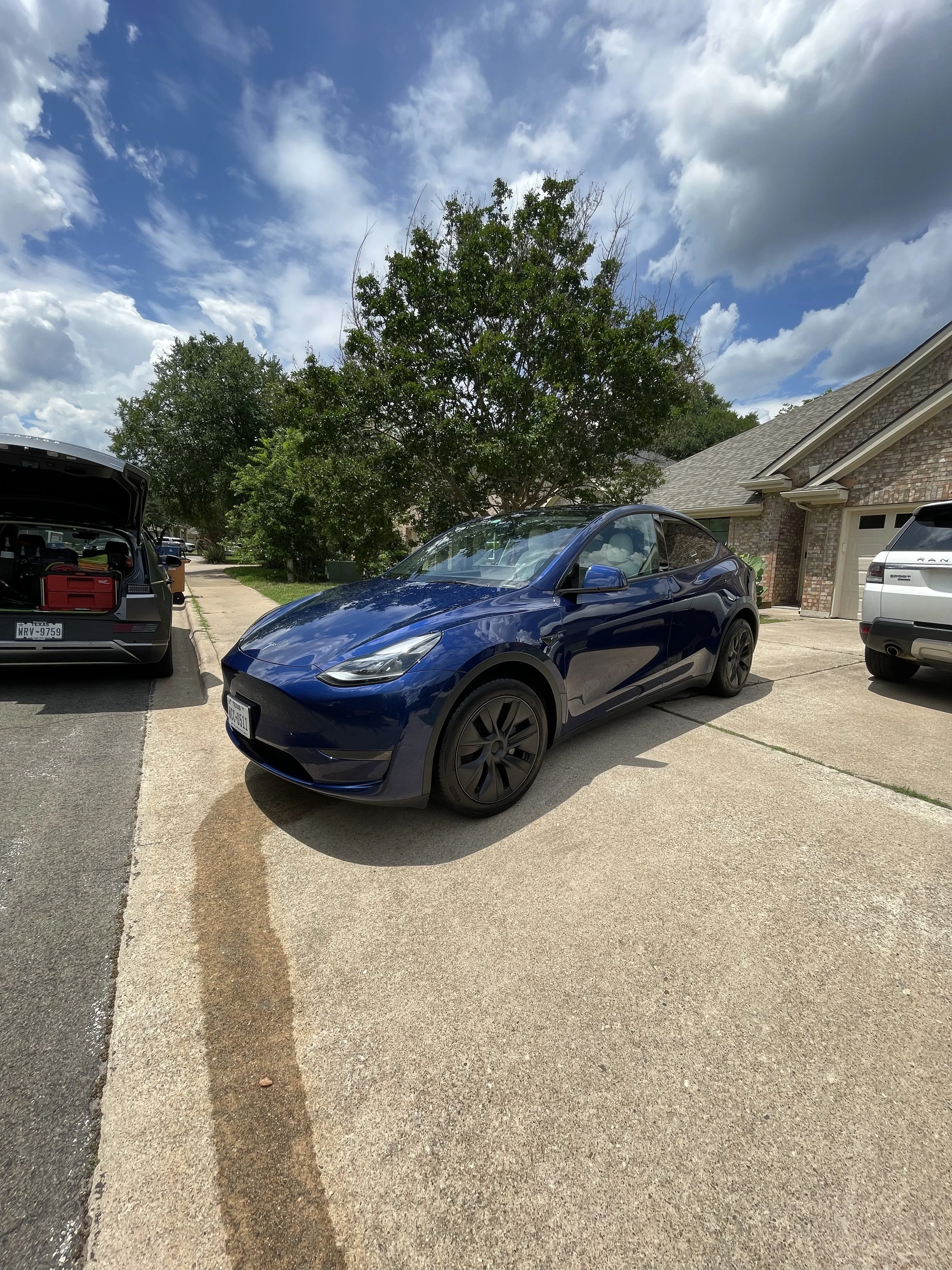 A residential driveway with several parked cars, a large tree, and a brick house. The sky is partly cloudy with blue patches visible.