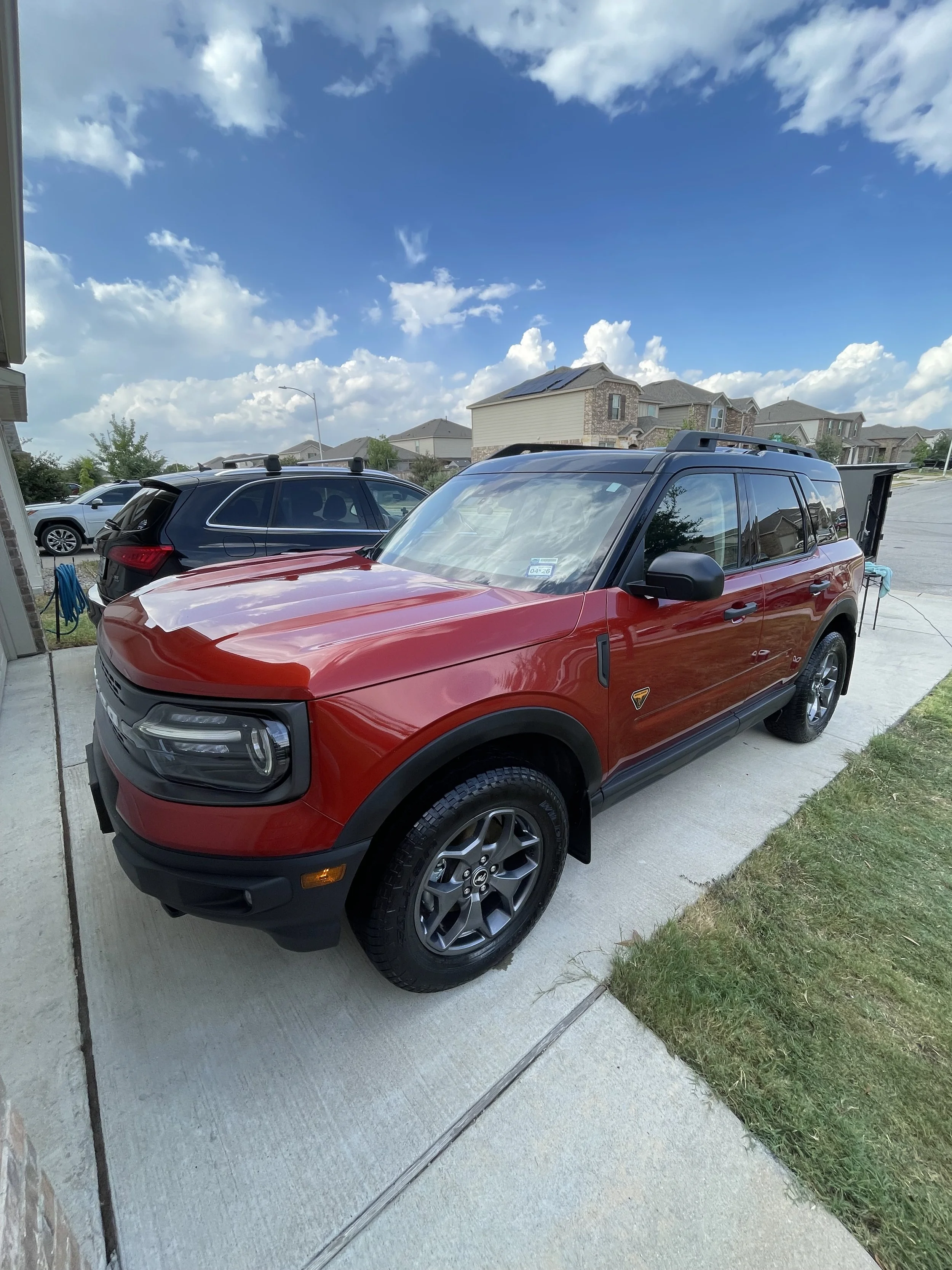 A red and black SUV parked on a concrete driveway in a suburban neighborhood with houses in the background and a partly cloudy sky.