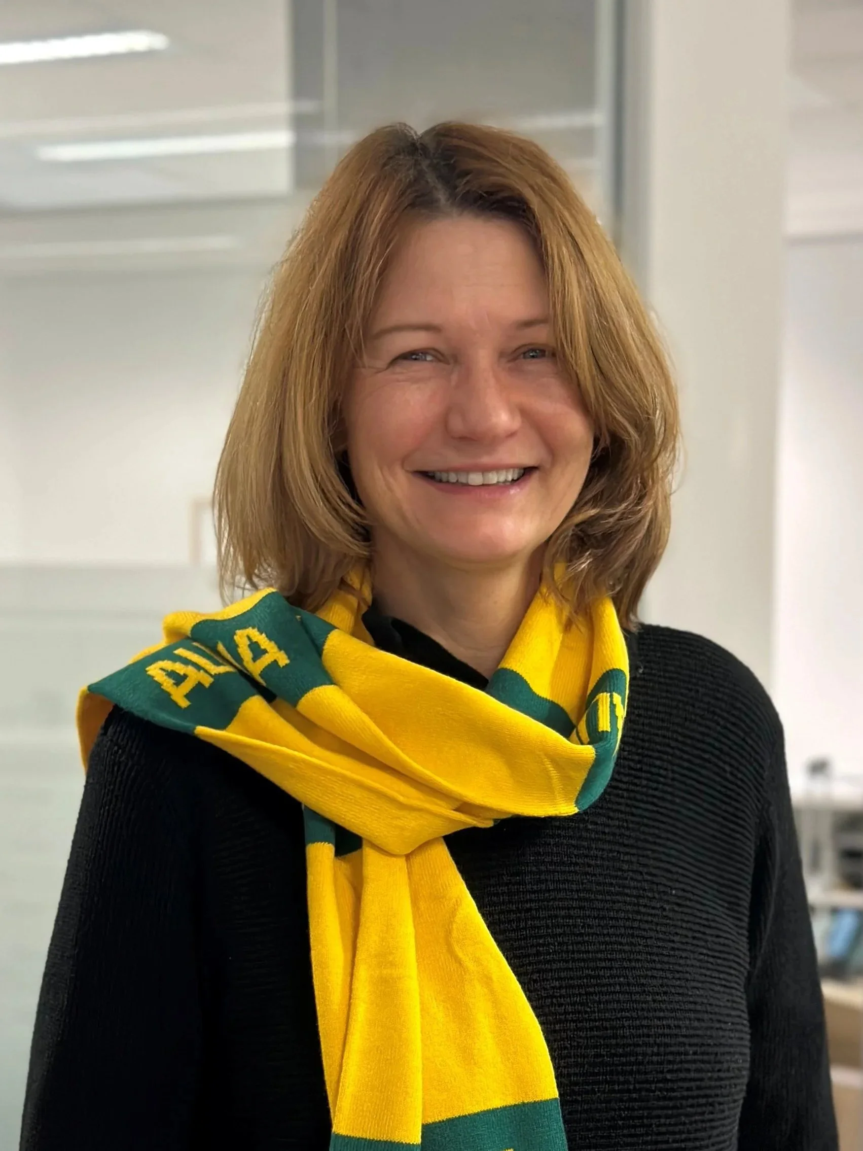 Woman with shoulder-length auburn hair smiling, wearing a yellow and green scarf and a black sweater, indoors.