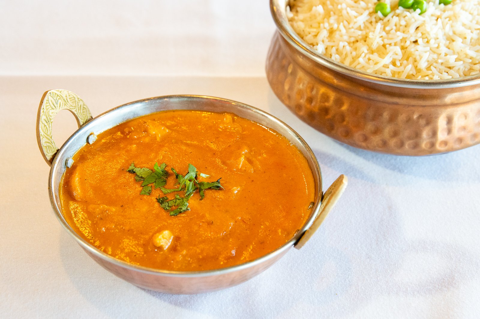 A bowl of Indian curry garnished with cilantro, and a bowl of cooked basmati rice with green peas on top, served on a white table.